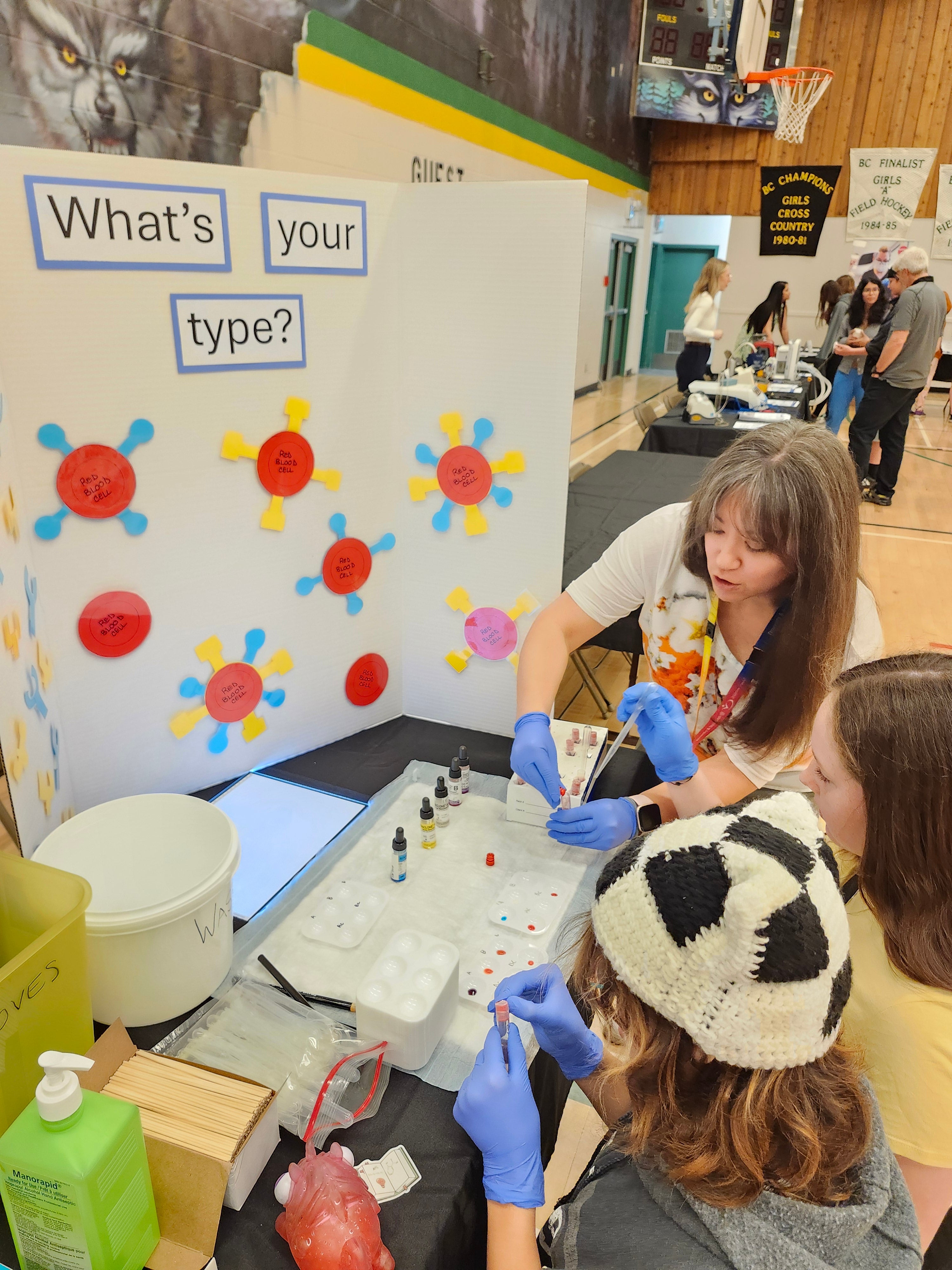 A woman with long grey blonde hair with blue medical gloves holds a test tube as two young people look on. The poster behind them says What's your type?