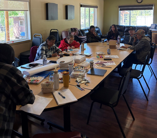 A group of people sits at a long table. Ink pens, paper and water bottles can be seen on the table.
