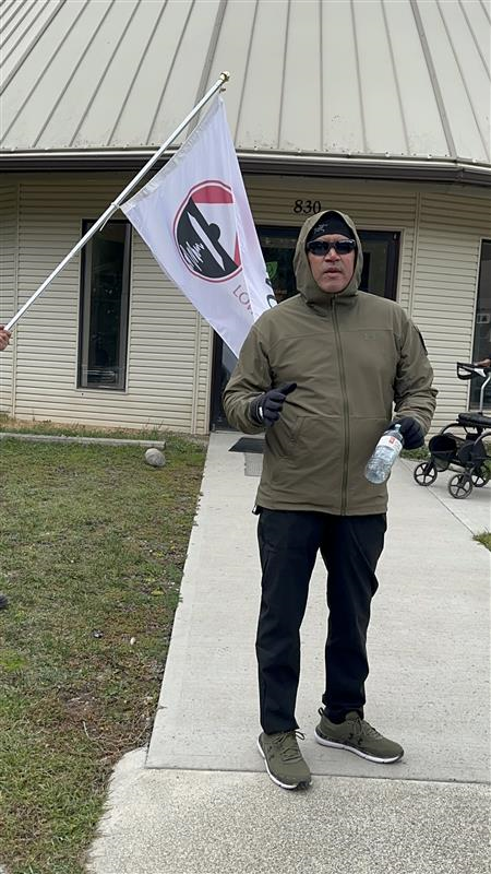 A person with a warm beige jacket wearing sunglasses next to a white flag gives a speech outside a building.