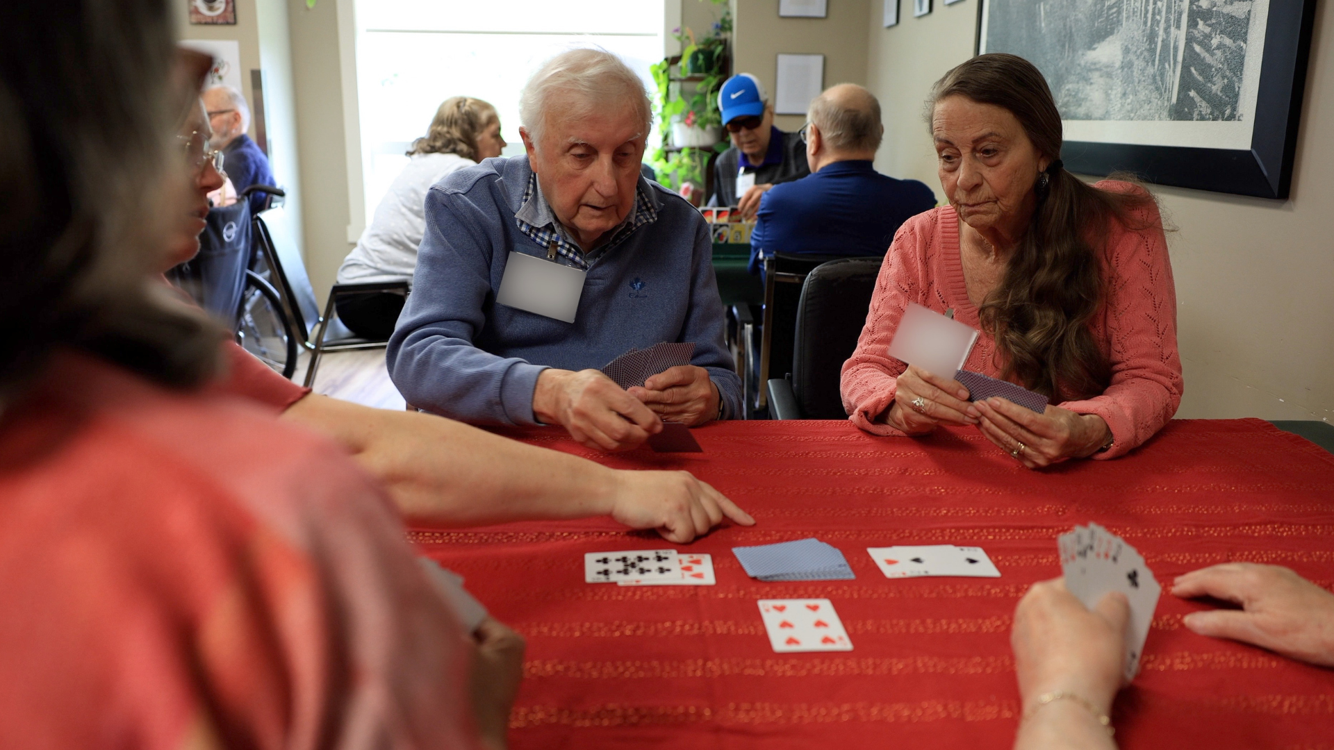 An older man and woman sit around a table with others playing cards.