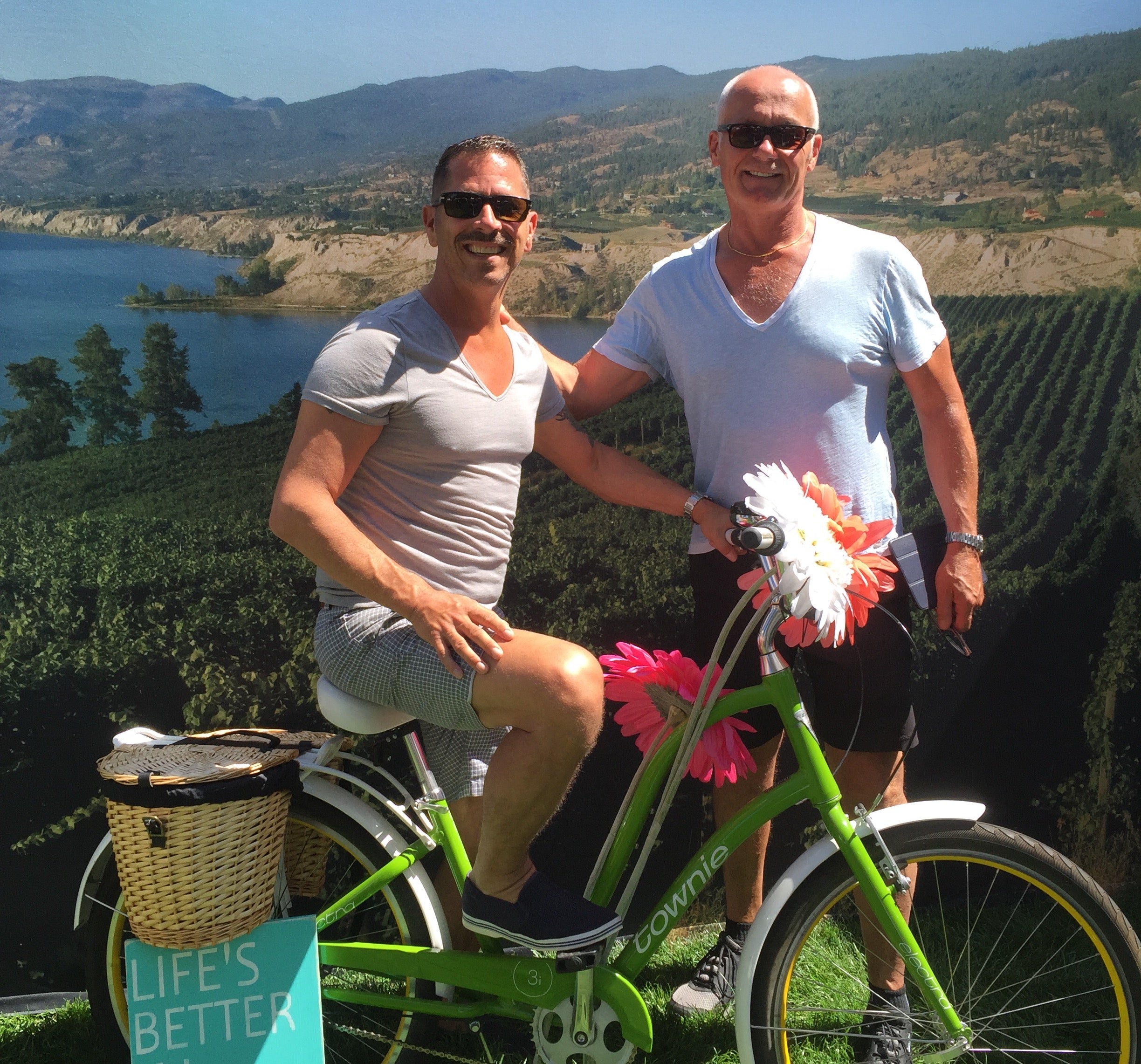 Two men in front of a vineyard and lake, one is sitting on a green bicycle covered in flowers