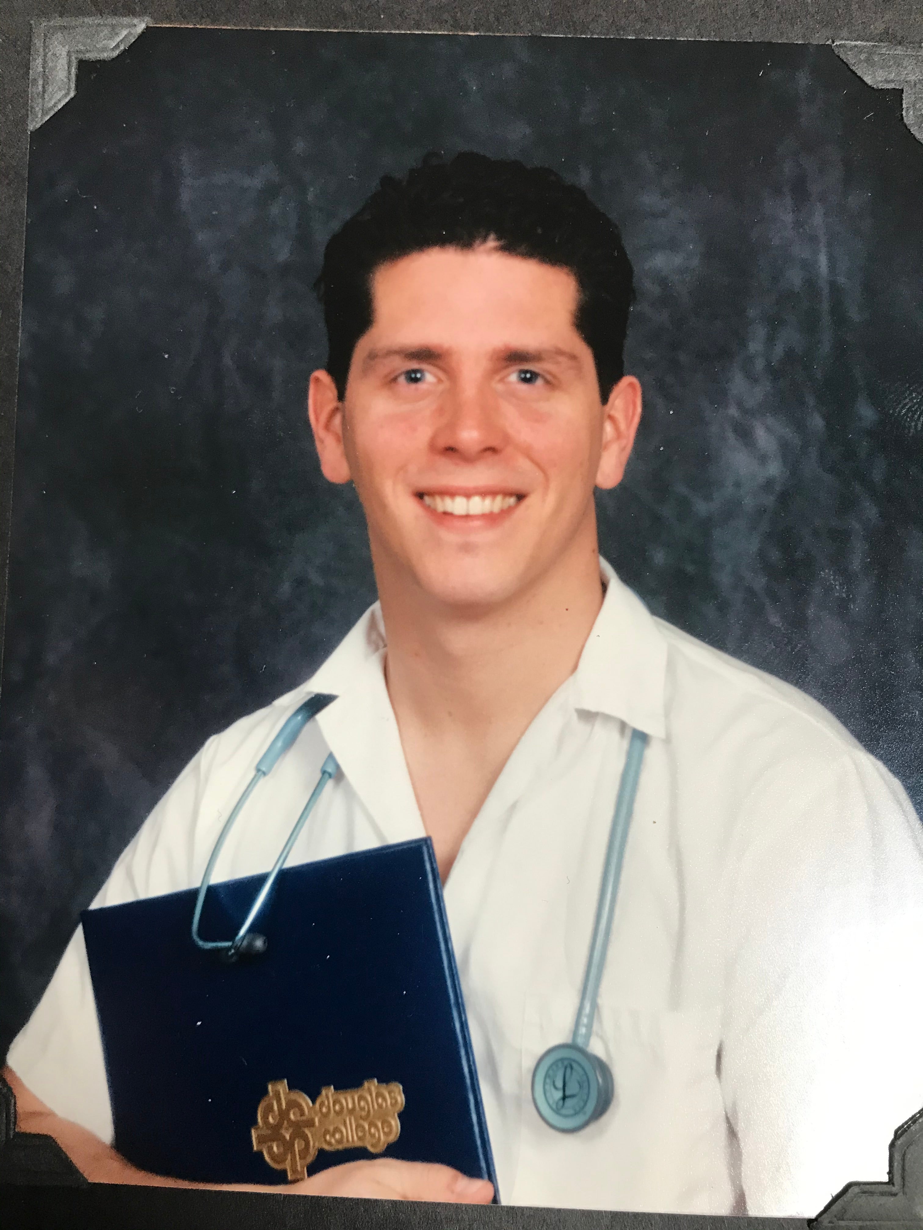 A man with short brown hair wearing a stethoscope and holding a degree certificate