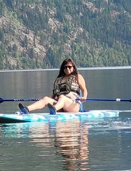 A person with long brown hair and sunglasses sits on a paddle board floating on the water while holding a long paddle.