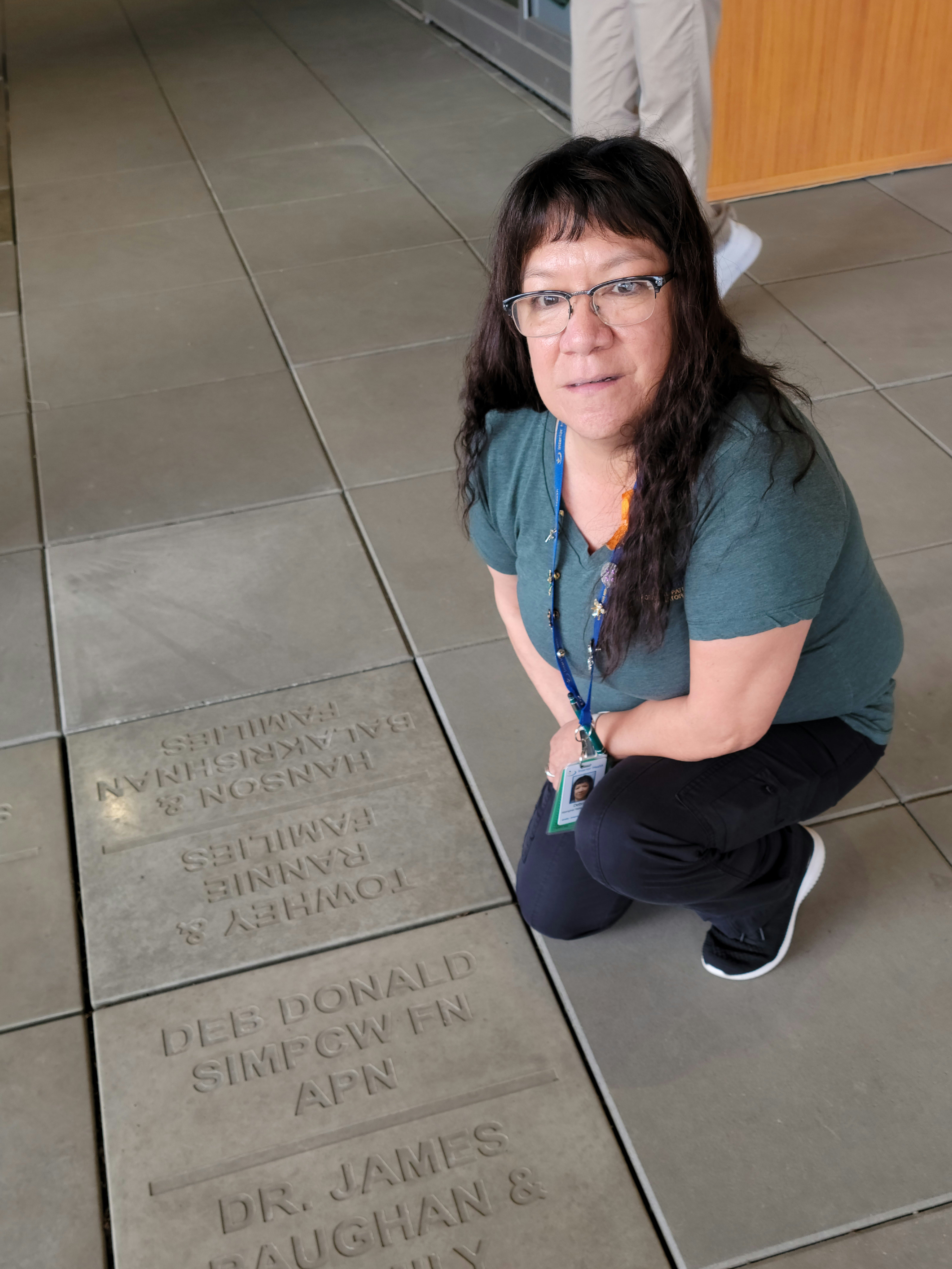 A casually dressed person with long brown hair and glasses kneels next to a floor with etched-in names.