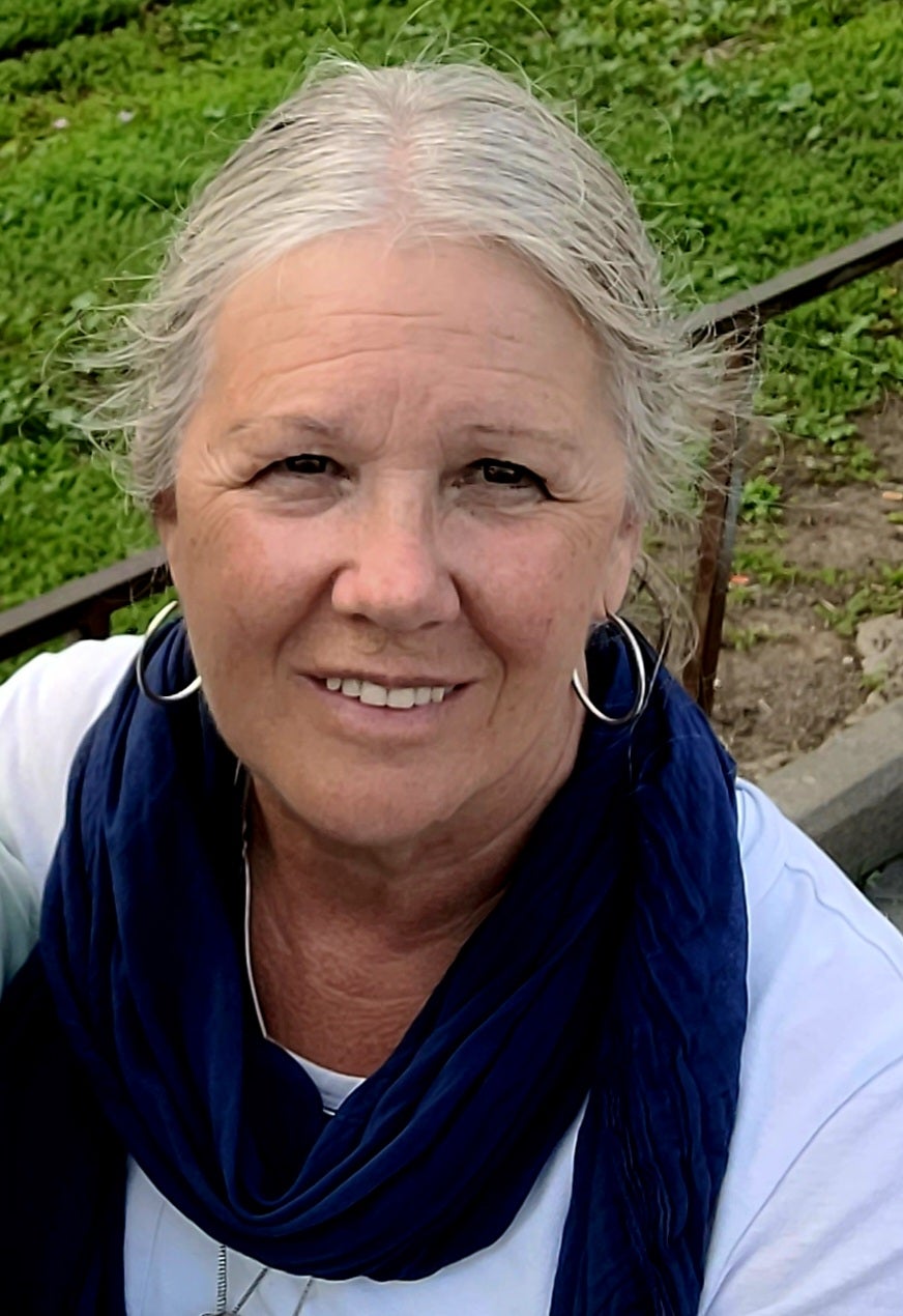 A smiling person with short silver hair wearing large hoop earrings, and a blue scarf wrapped around a white shirt.