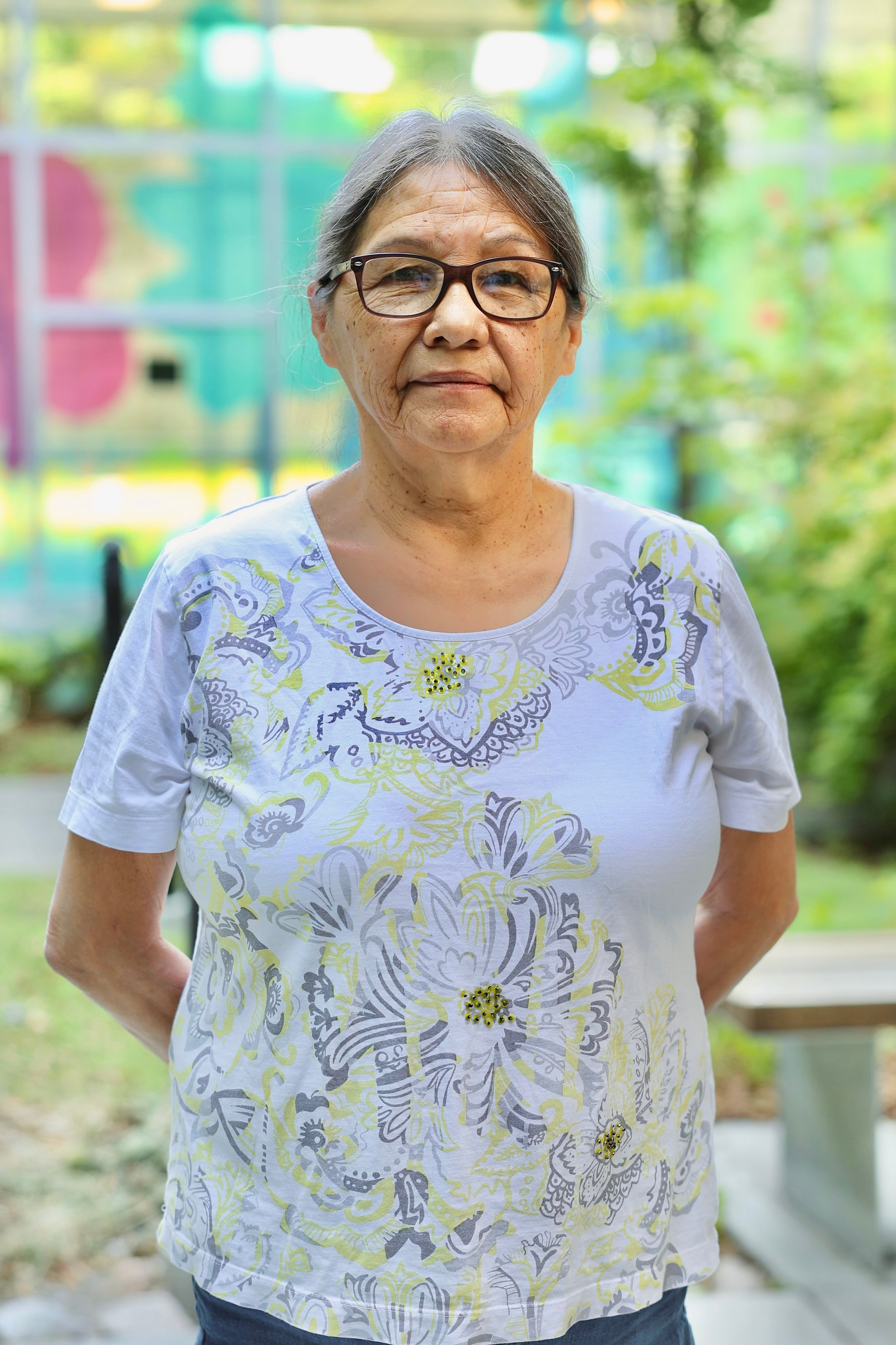 A middle aged person with silver brownish hair and black glasses, who is wearing a floral shirt.