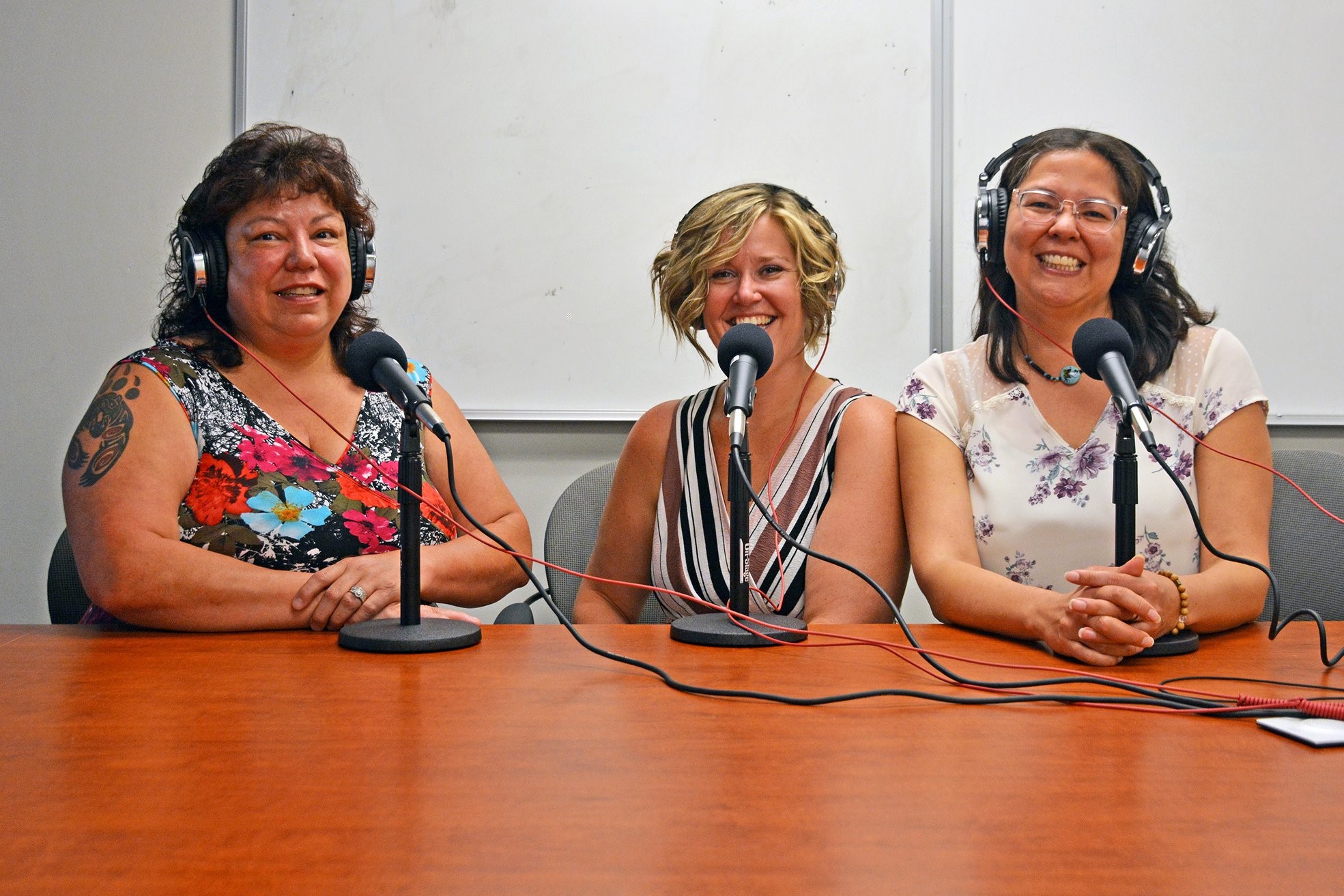 Three women sitting in a recording studio with headphones on and microphones in front of them pose for a picture in a recording studio.