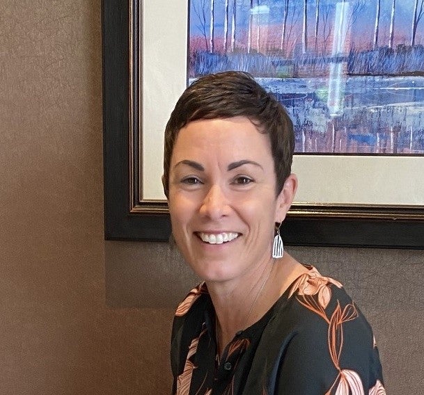 A woman with short brown hair and long white earrings wearing a green blouse sits in a room painted brown. There's a blue painting behind her.
