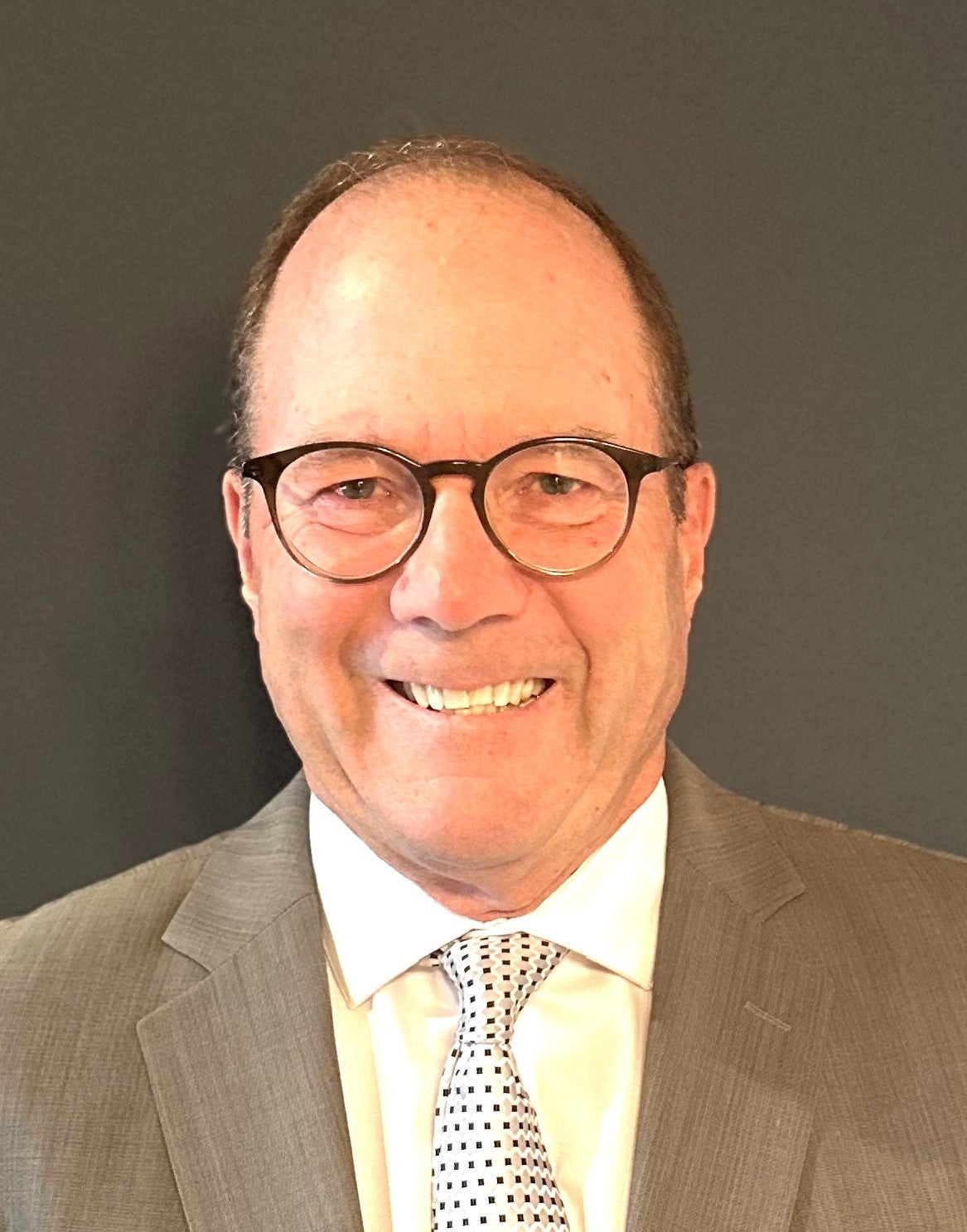A smiling person with short brown hair wearing glasses and a business suit and tie.