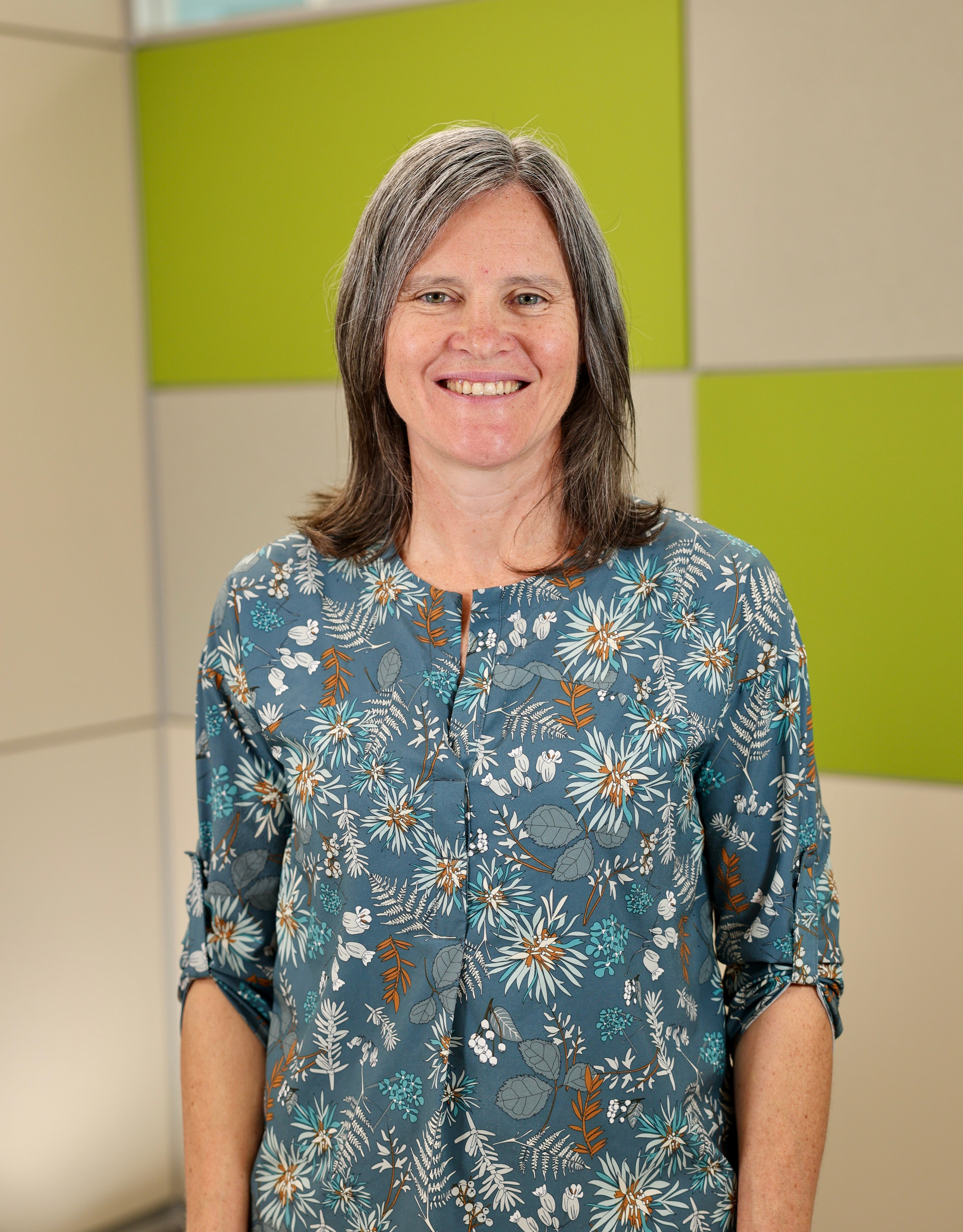 A head and shoulders photo of a smiling person with medium length grey hair wearing a green flowered blouse standing in front of a wall with beige and green squares