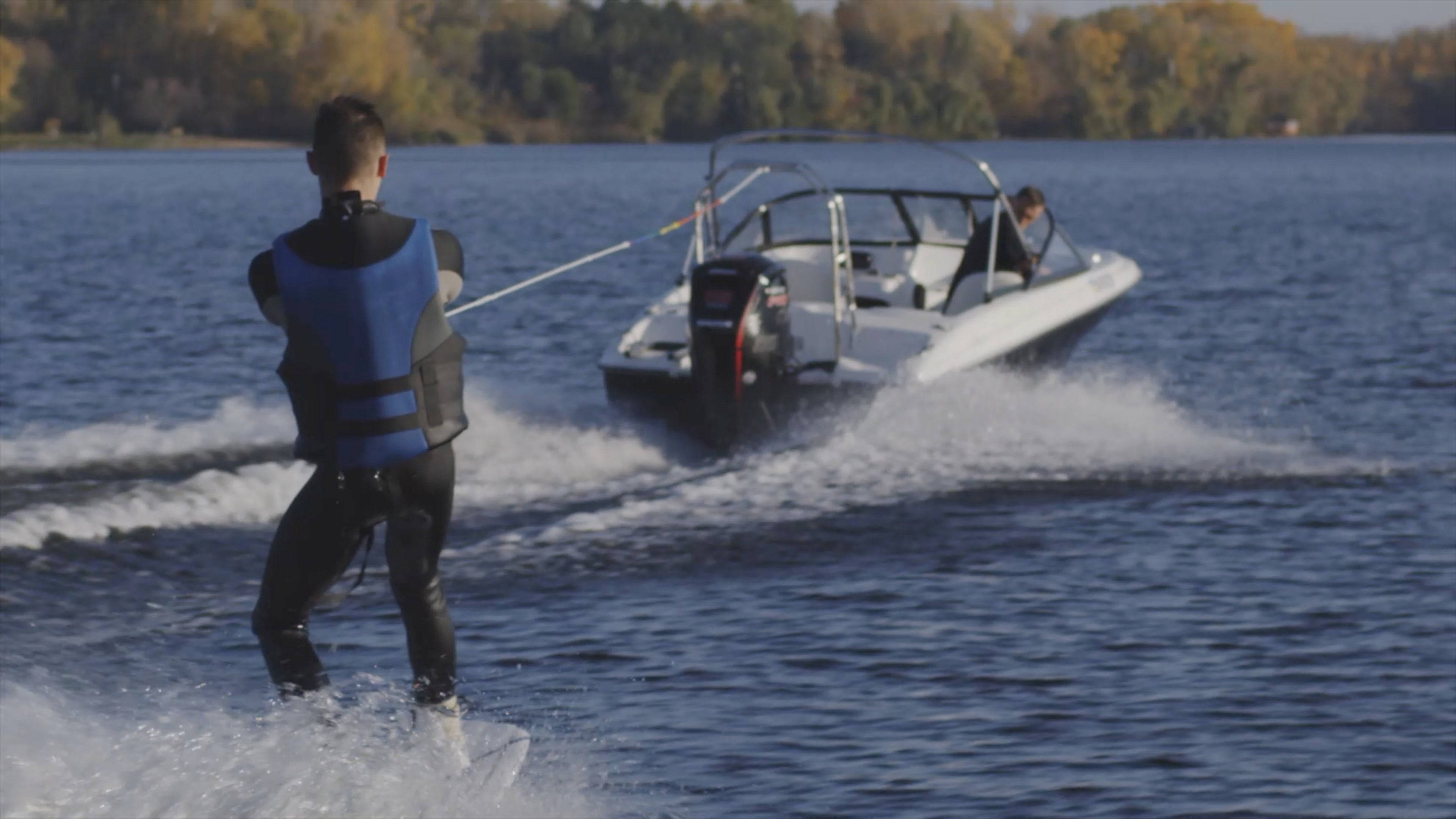 A person wearing a lifejacket on a wakeboard is pulled behind a boat on a lake on a sunny day.