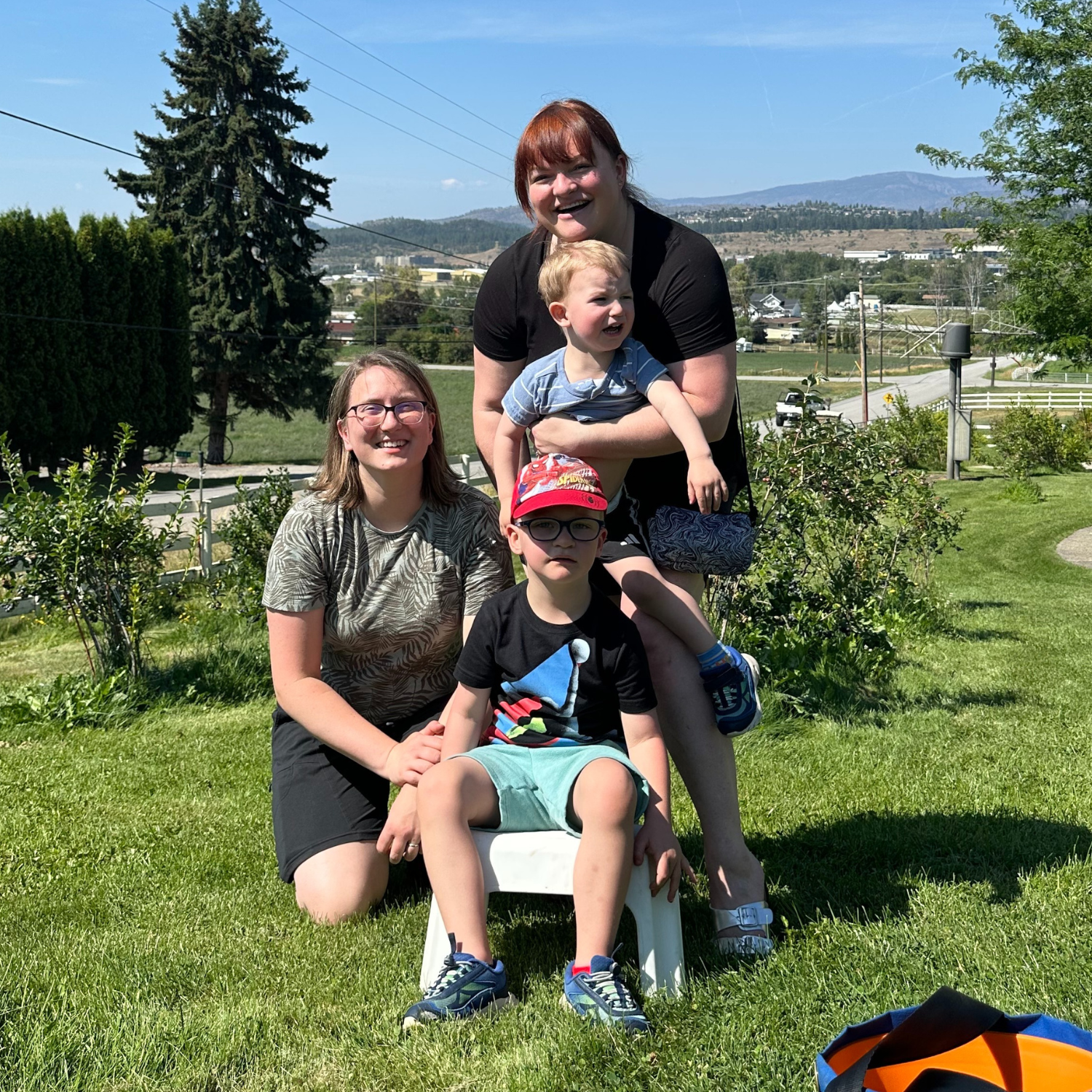 A woman kneeling on grass and another standing up, holding her young son. Her other son is sitting in front of her on a stool.