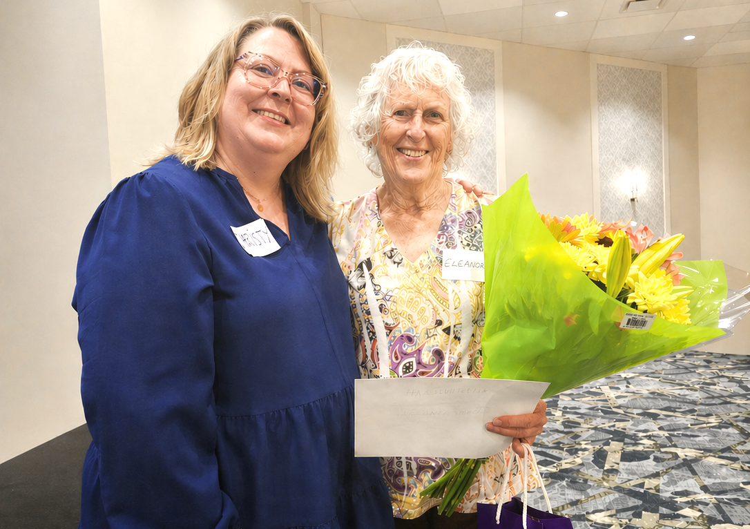 A person wearing glasses and a blue shirt stands next to an older person holding a bouquet of yellow flowers.