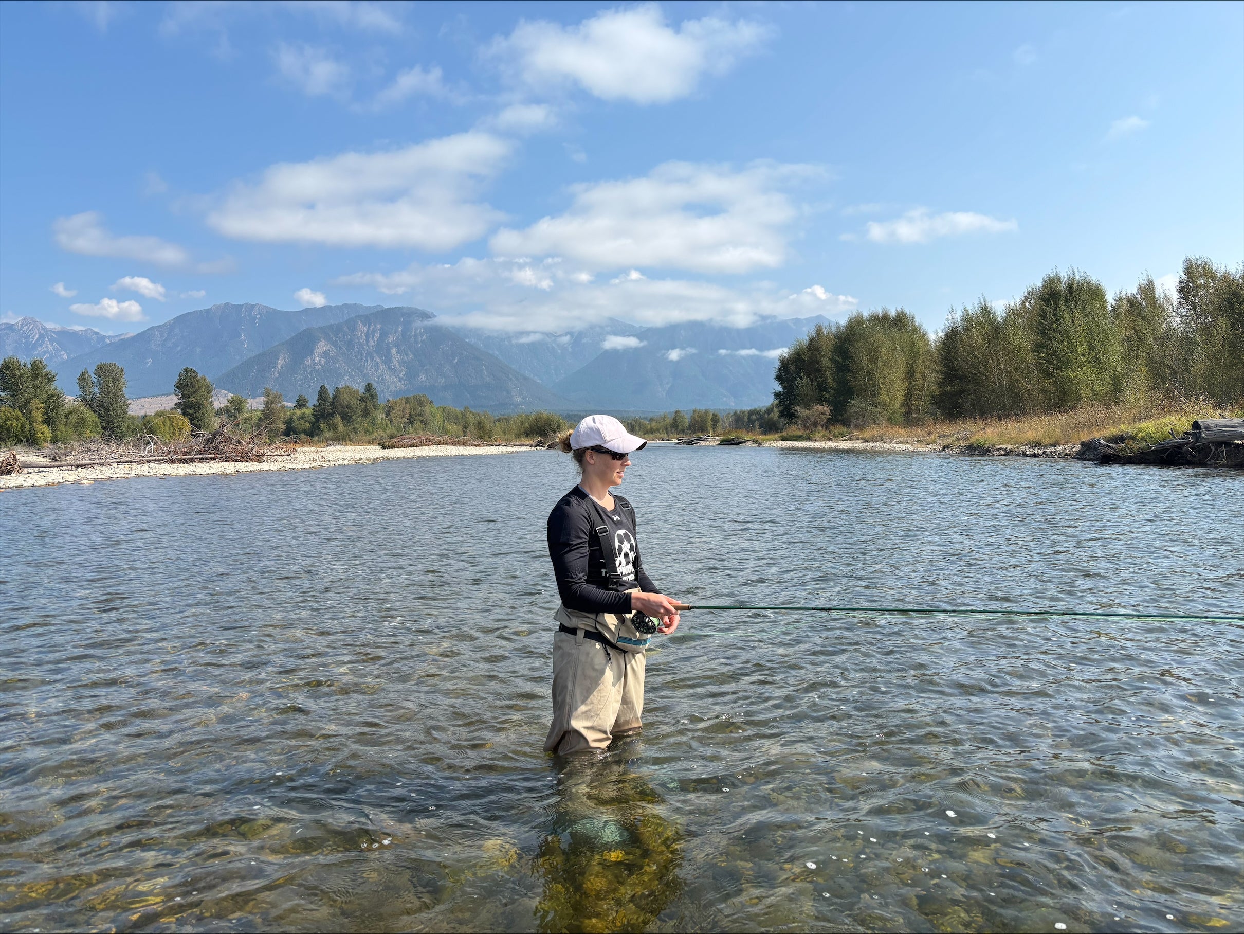 A person in a white ball cap, black shirt and khaki waders stands in a clear river on a sunny day casting a fishing line.