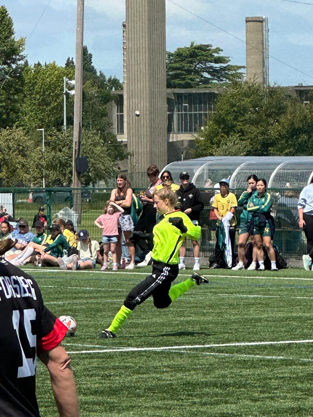 A person in a neon yellow jersey is set to strike a soccer ball on a grass pitch.