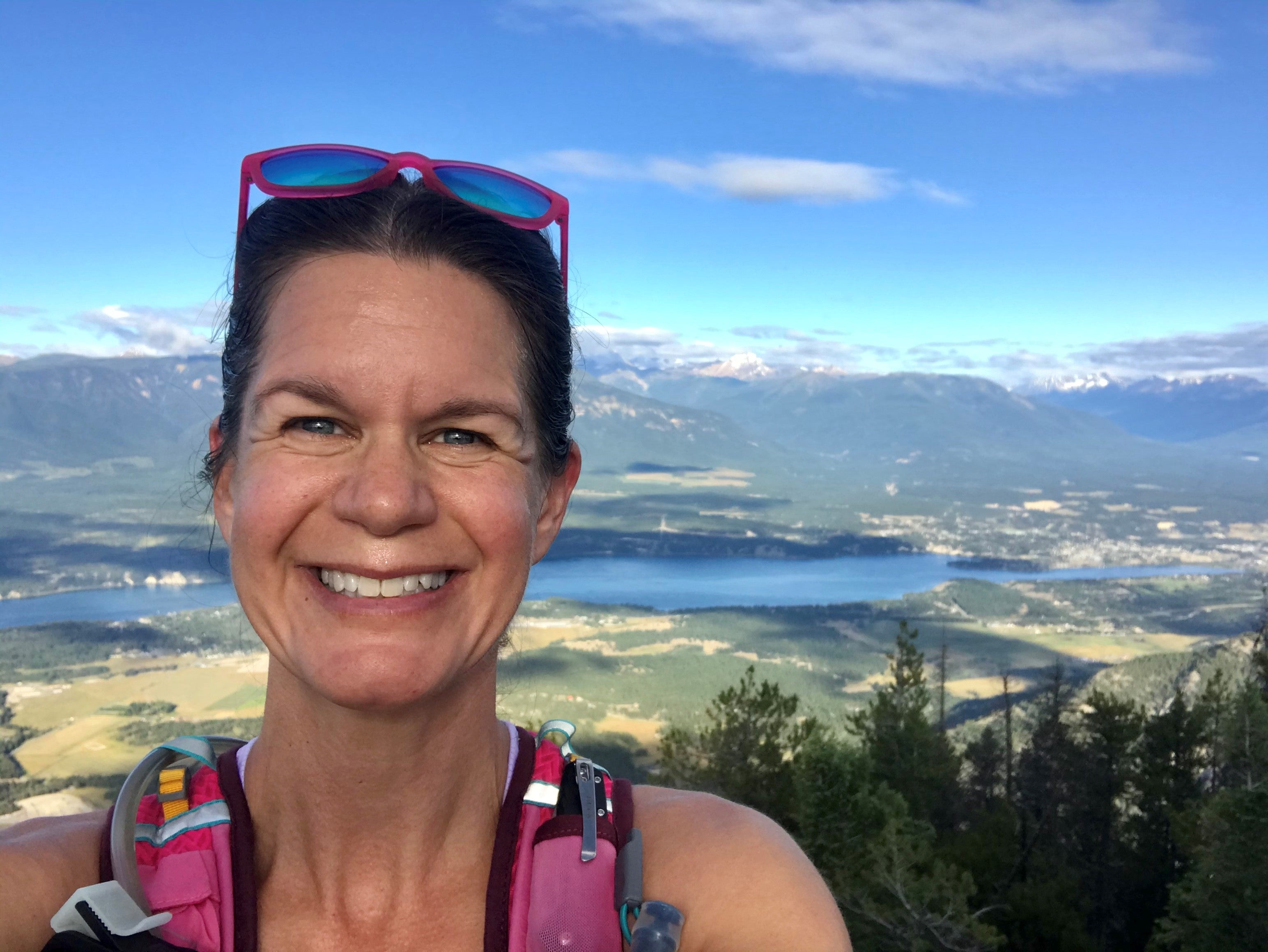 Woman standing in front of mountains and a lake, smiling, with sunglasses on her head.