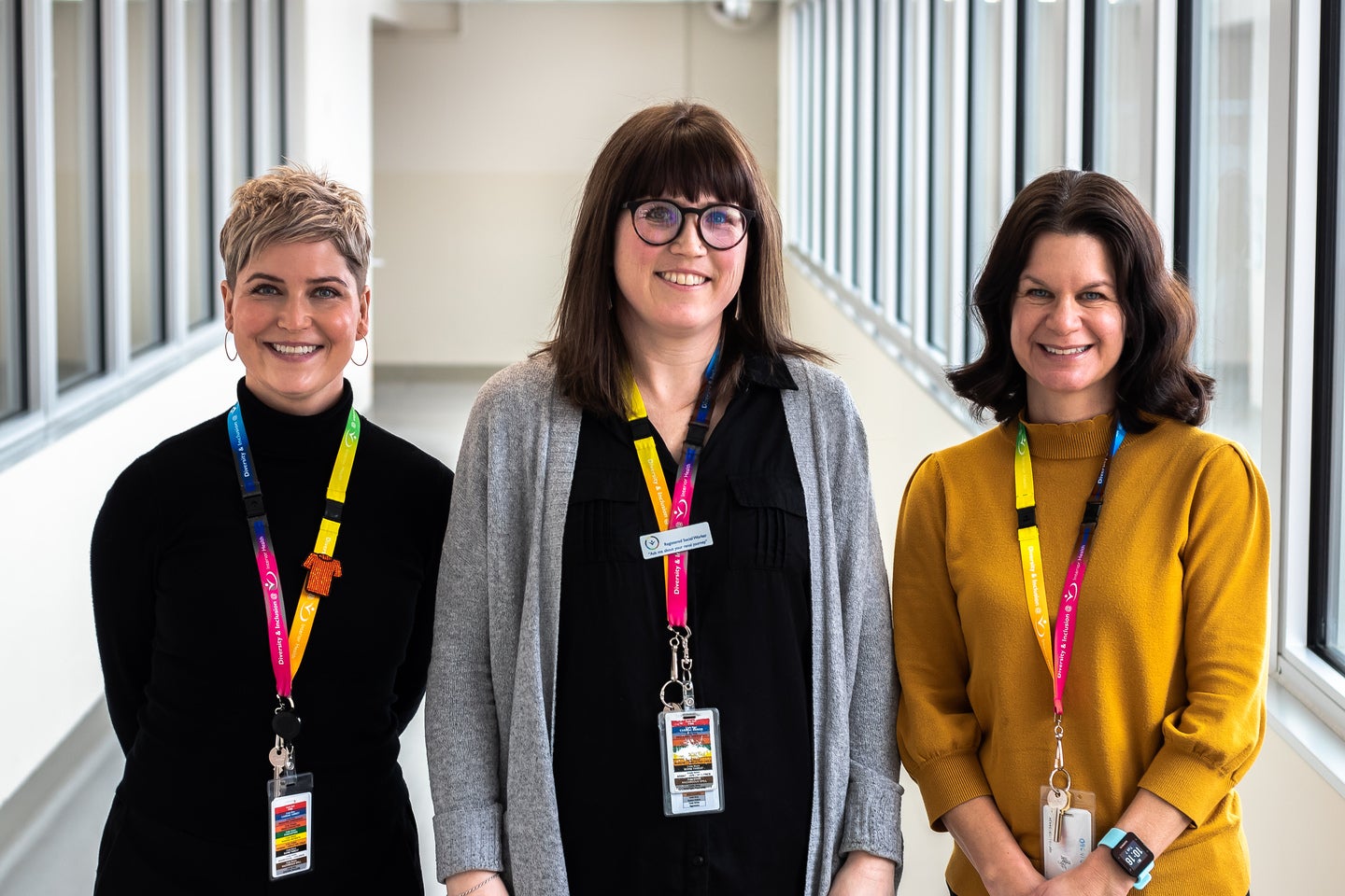 Three women standing next to each other. The first (left) is wearing black and has short, blonde hair. The second (center) has brown hair, bangs, and is wearing glasses, and the third (right) wears a yellow turtleneck and has brown hair.