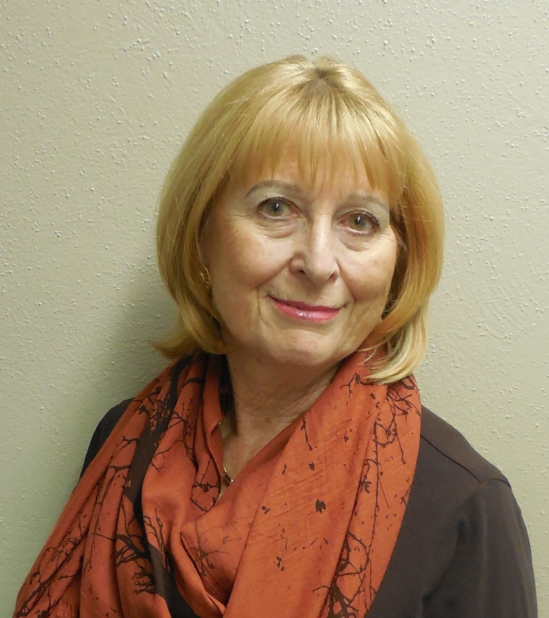 A person with strawberry blonde hair wearing a loose orange scarf and brown sweater in front of a beige wall.