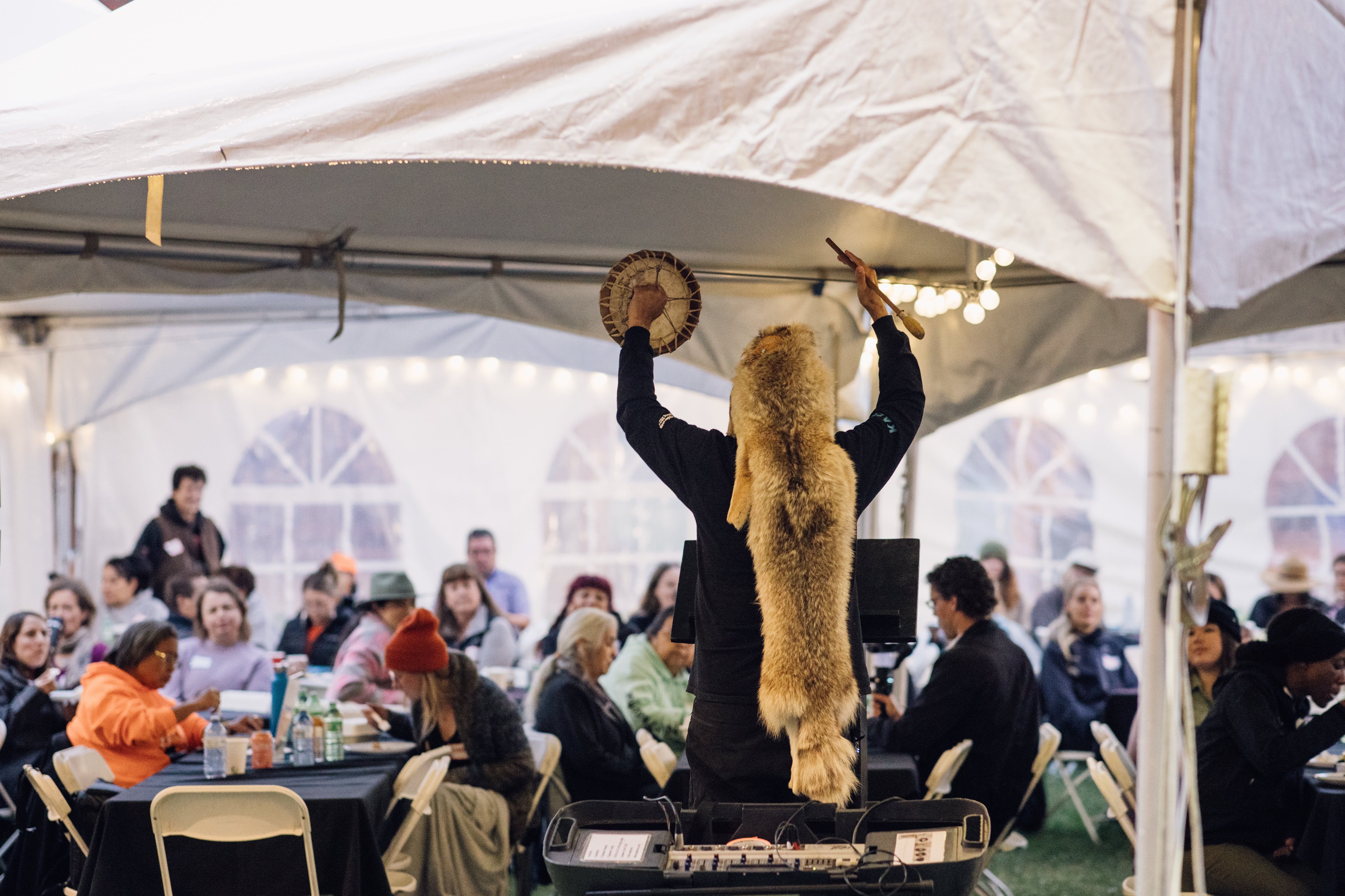 People are sitting at tables underneath a white tent watching a performer.