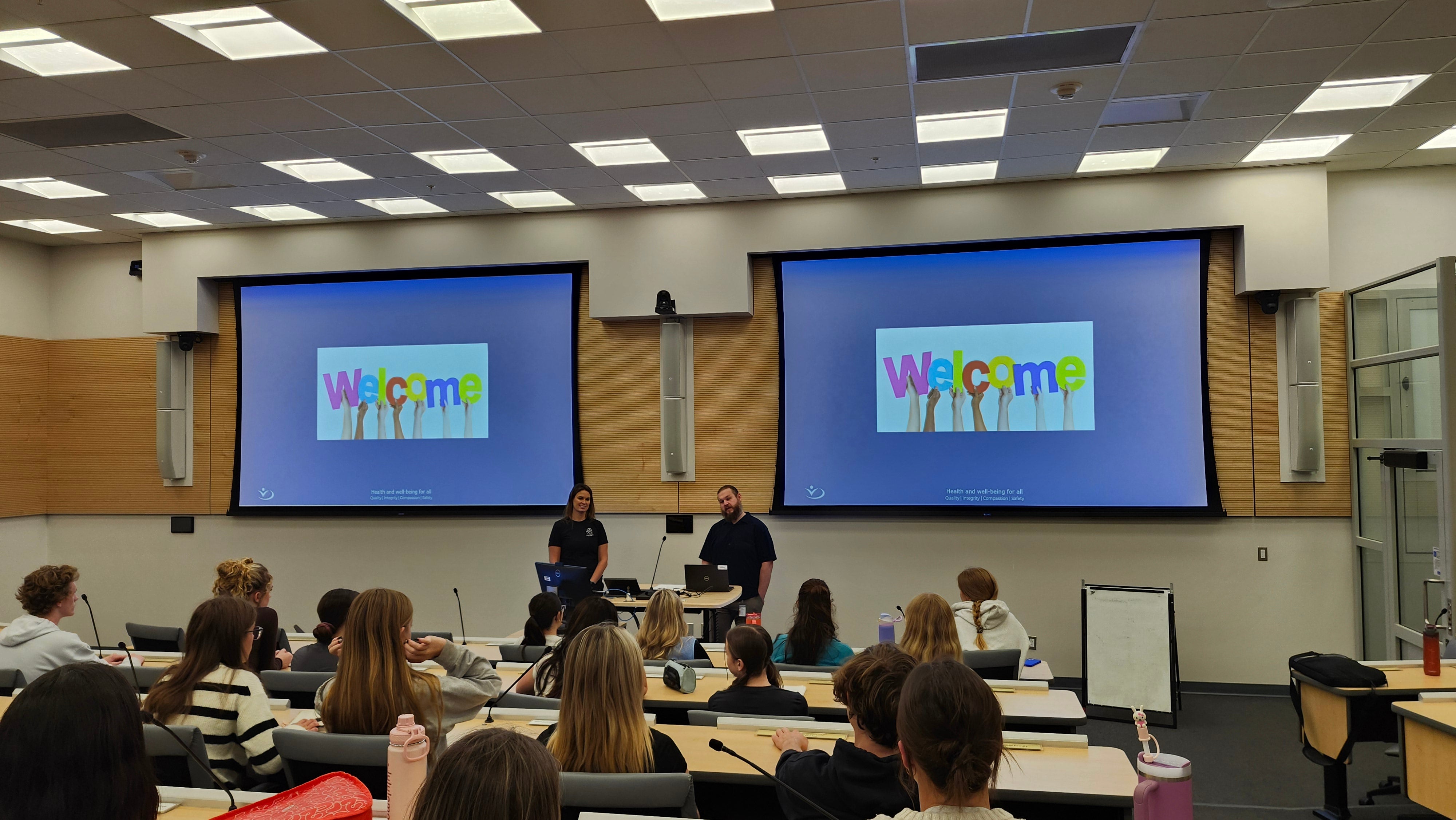 Two people stand at the front of a lecture theatre at a lectern with two big screens above them that both say welcome.