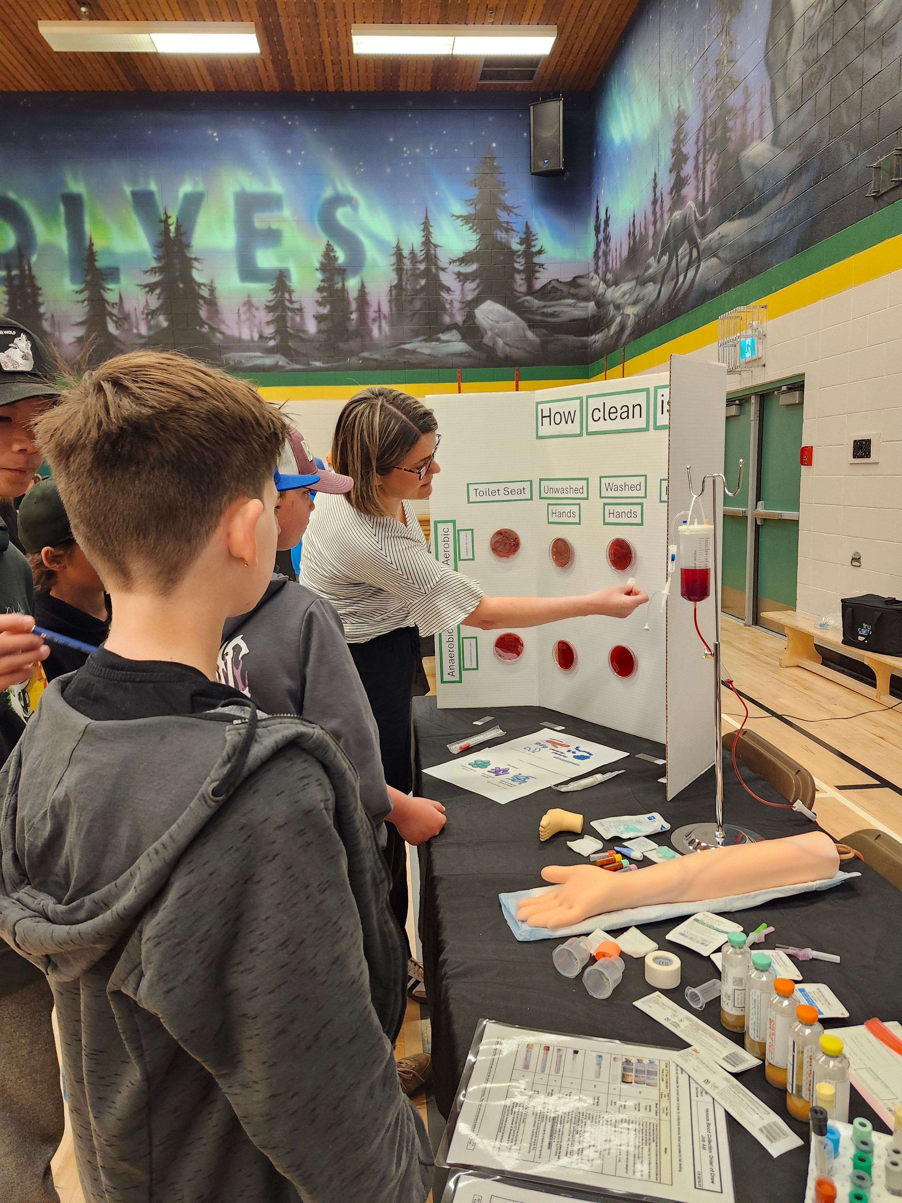 Young students watch on as a woman in a white striped blouse points to petri dishes posted on a poster board. The table in front shows various bottles and mannequin arm.