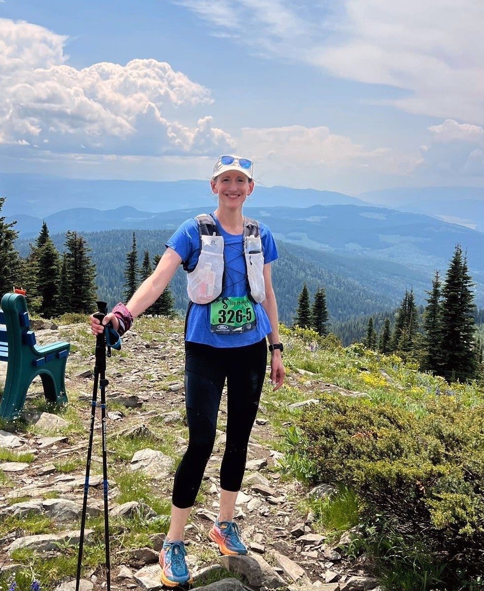 Participant wearing a race bib numbered 3262, standing on a rocky trail with pine trees and mountains in the background, under a clear sky with clouds.