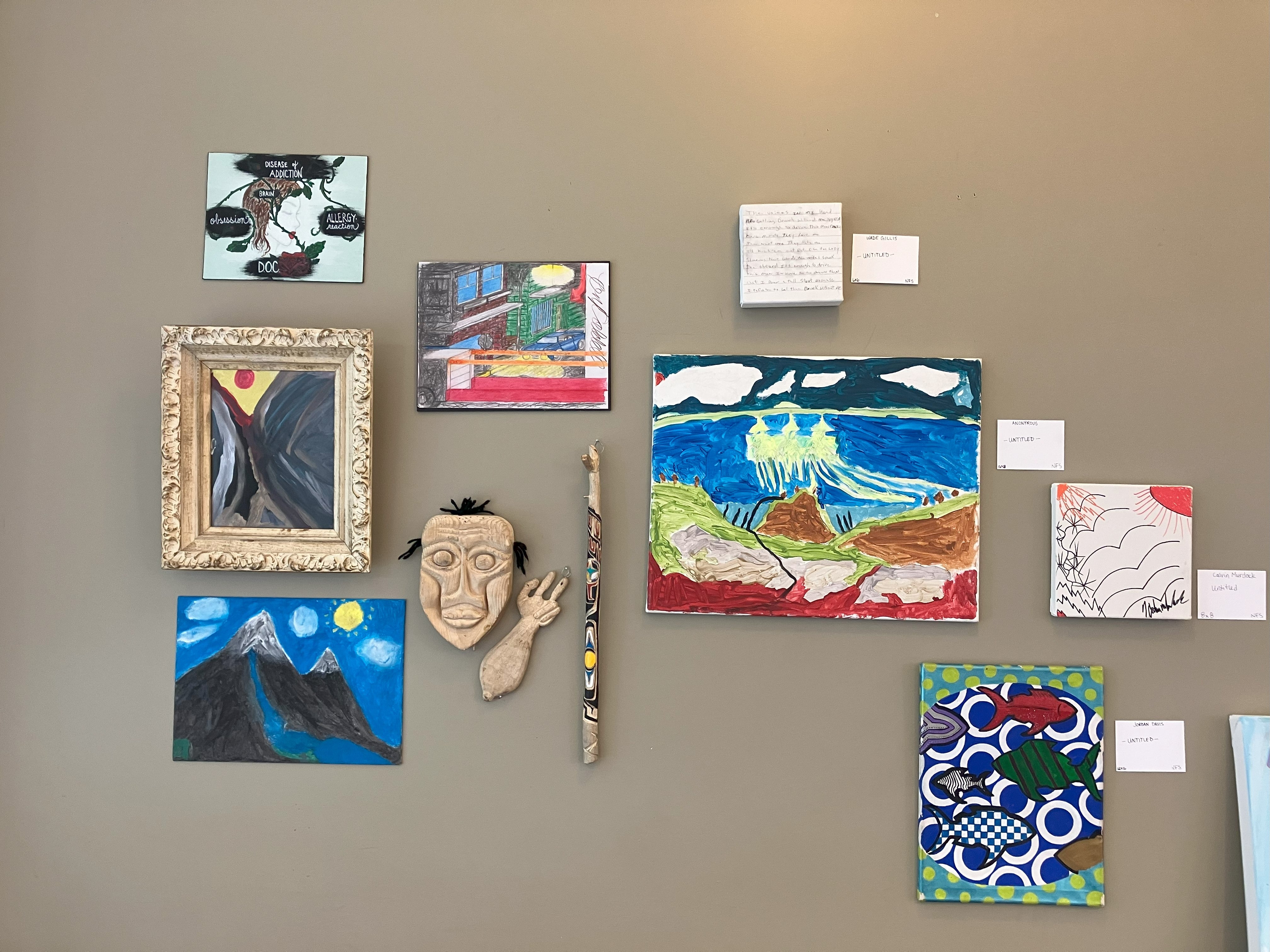 A display of art on a beige wall. Some of the art is unframed and includes a mask and talking stick.