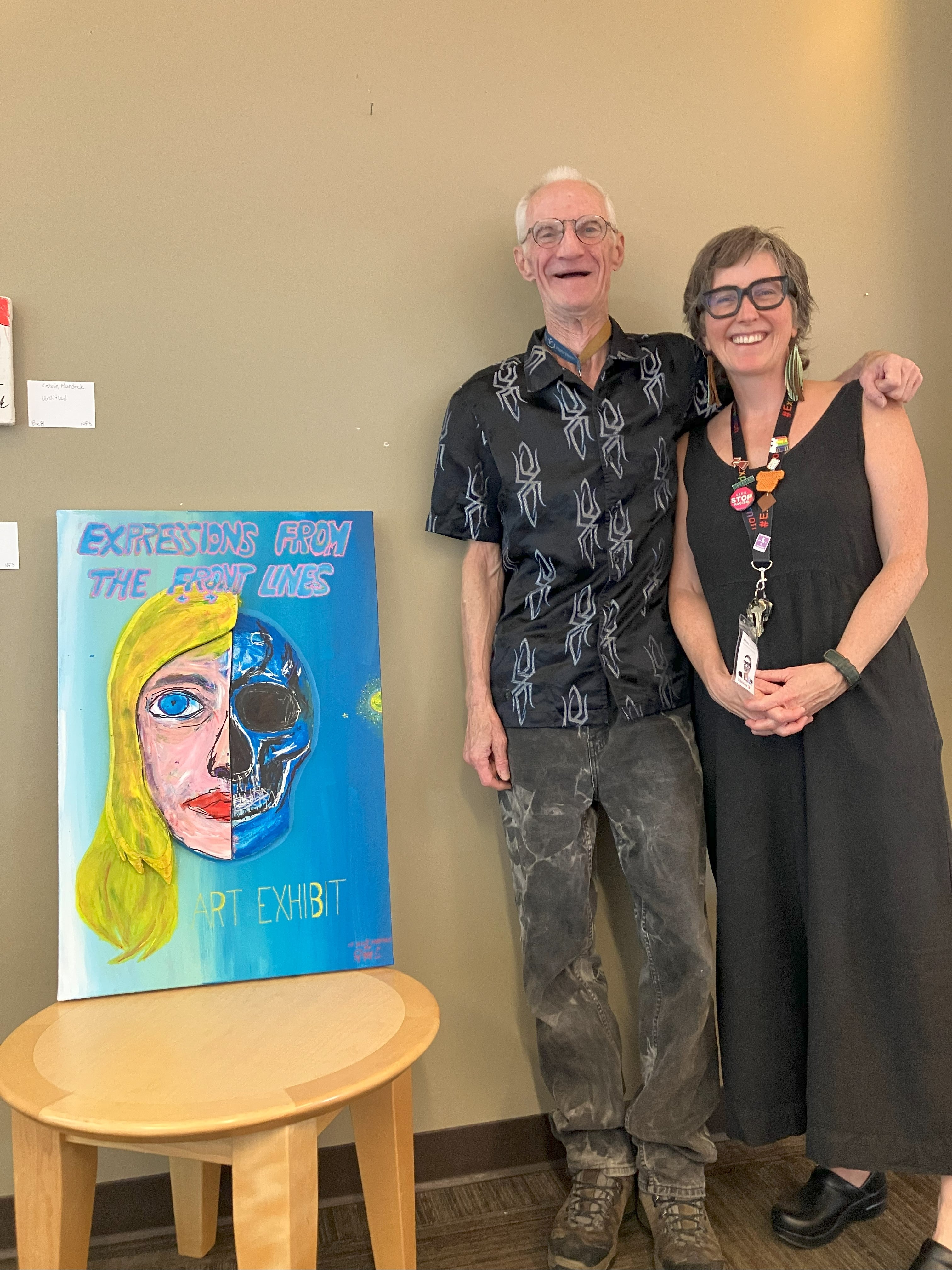 Two people stand side by side next to a painting on a small table that shows half a woman's face and half a skull.