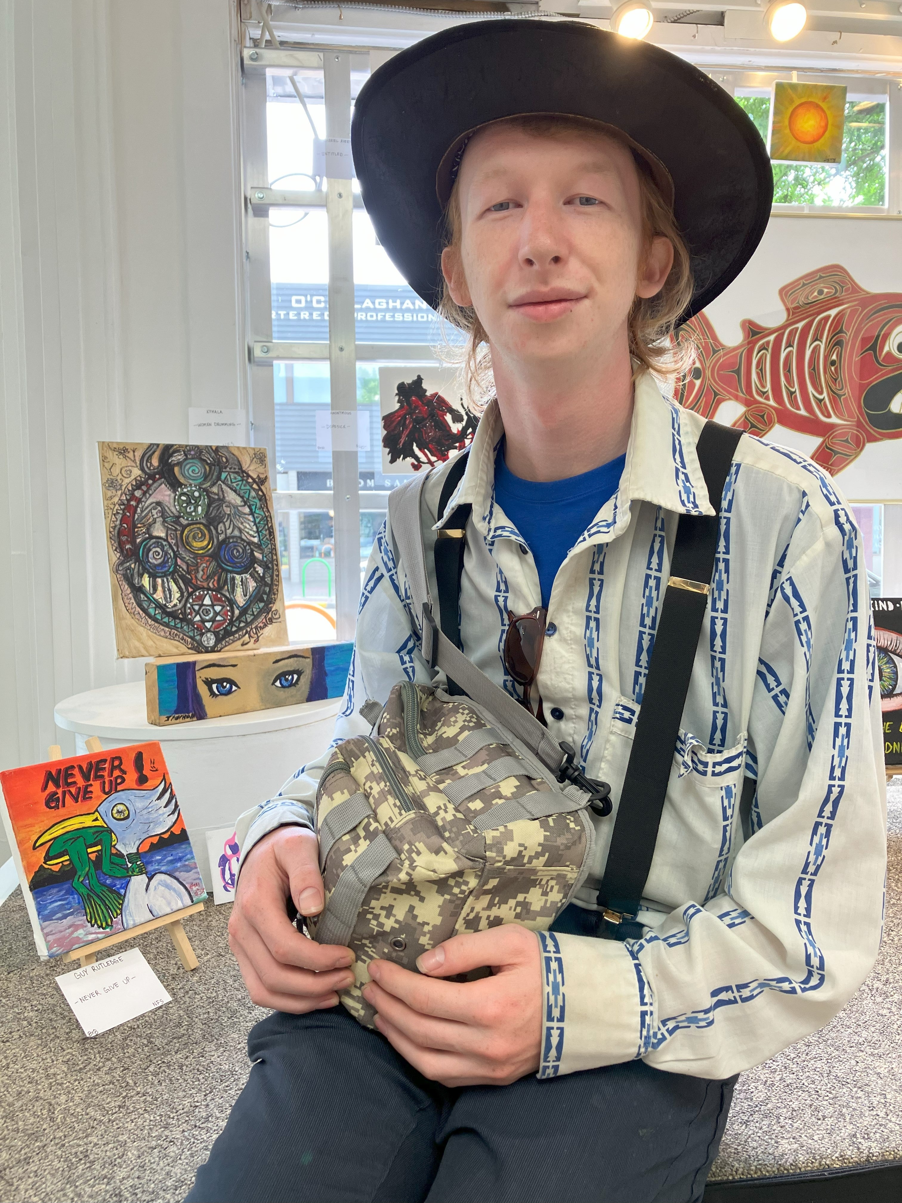 A person wearing a wide-brimmed black hat and striped shirt and suspenders sits in front of a window art display.