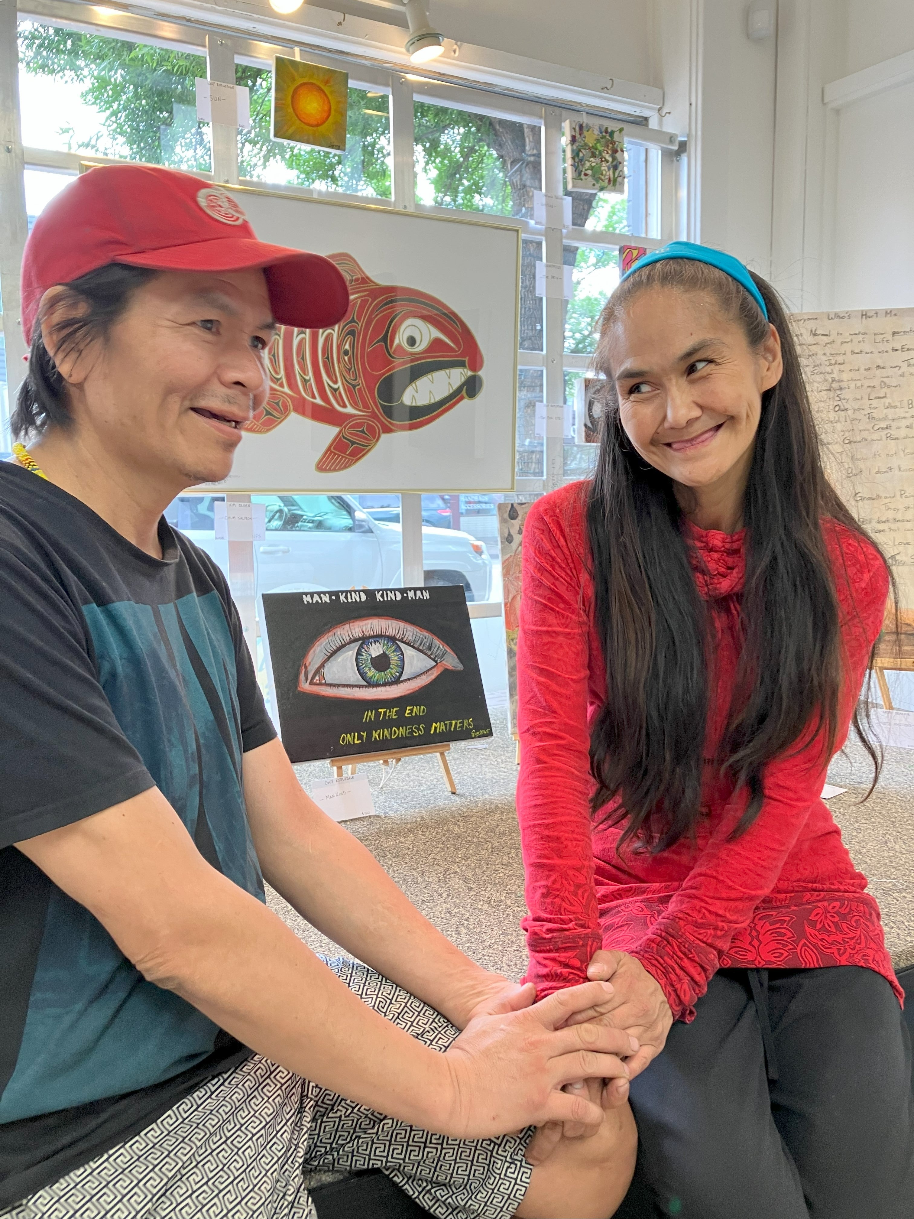 Two people sit beside each other holding hands. One is wearing a ball cap and blue tshirt and the other has long black hair and is wearing a long-sleeved red shirt.