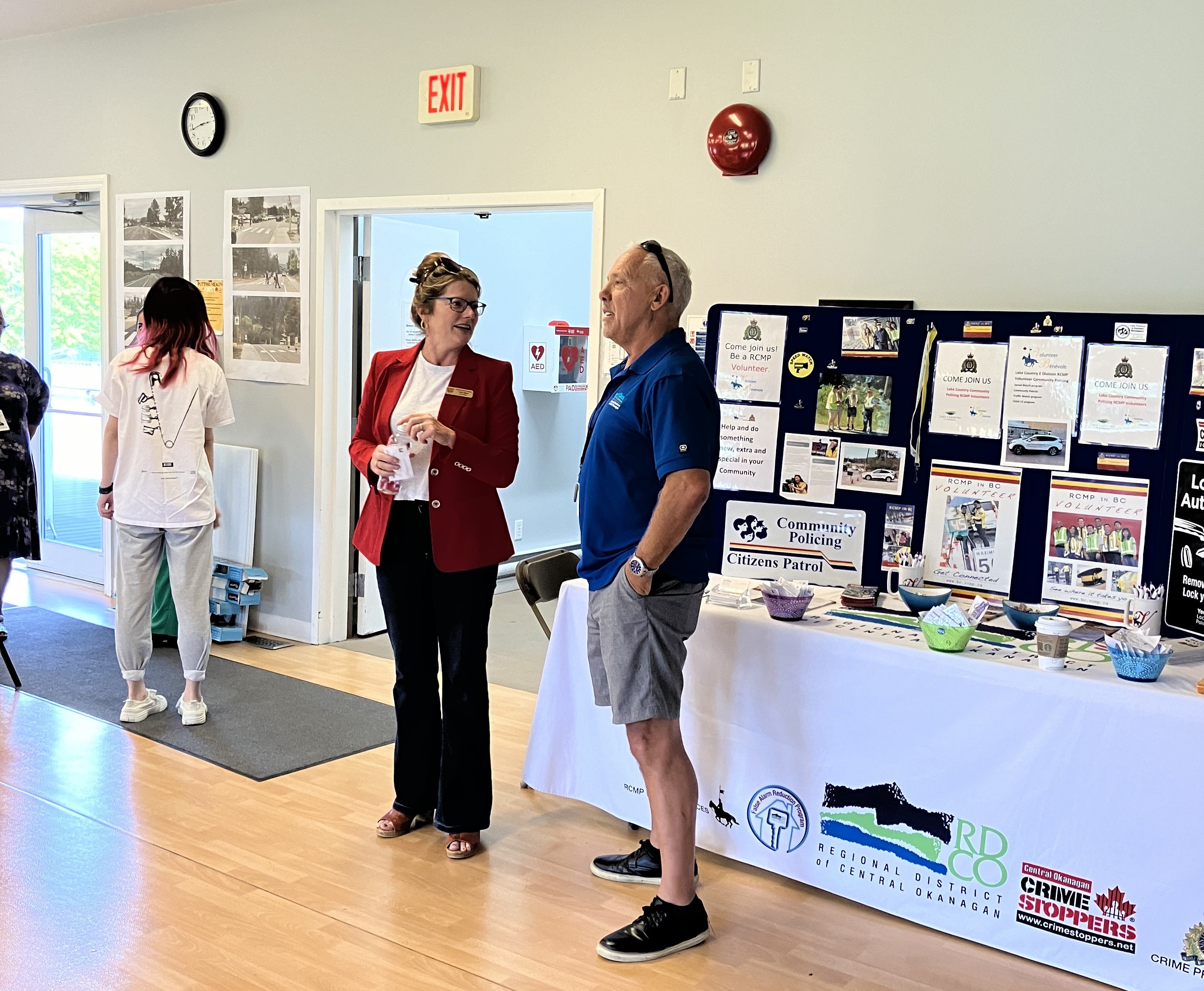 A woman in a red business jacket and black pants with her hair tied back and glasses on her head chats amicably with a man wearing a blue polo shirt and grey shorts. They stand in front of a poster display with a sign that says Community Policing and Citizens Patrol.