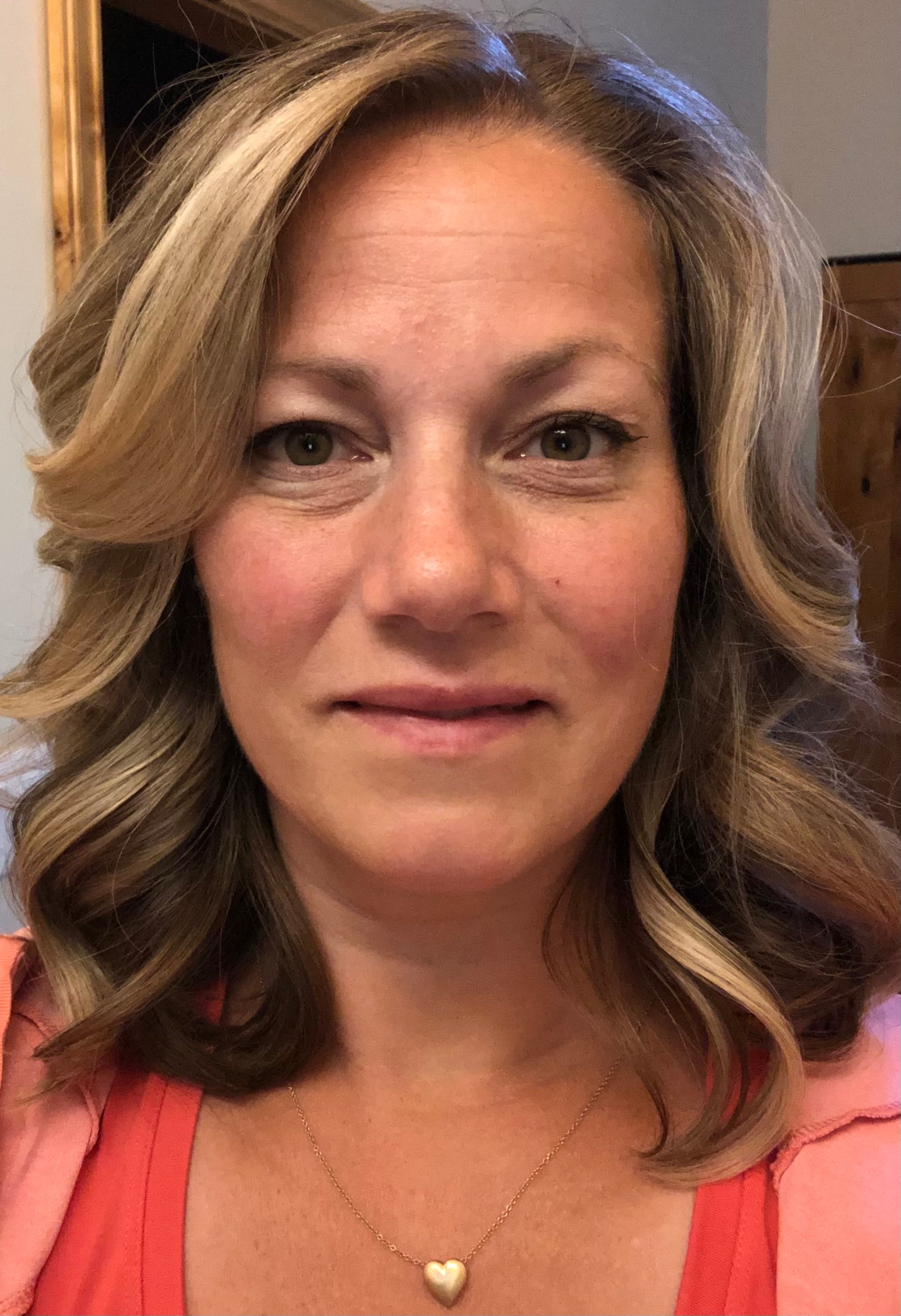 A person with wavy blonde hair framing their face wearing an orange tank top and silver necklace.