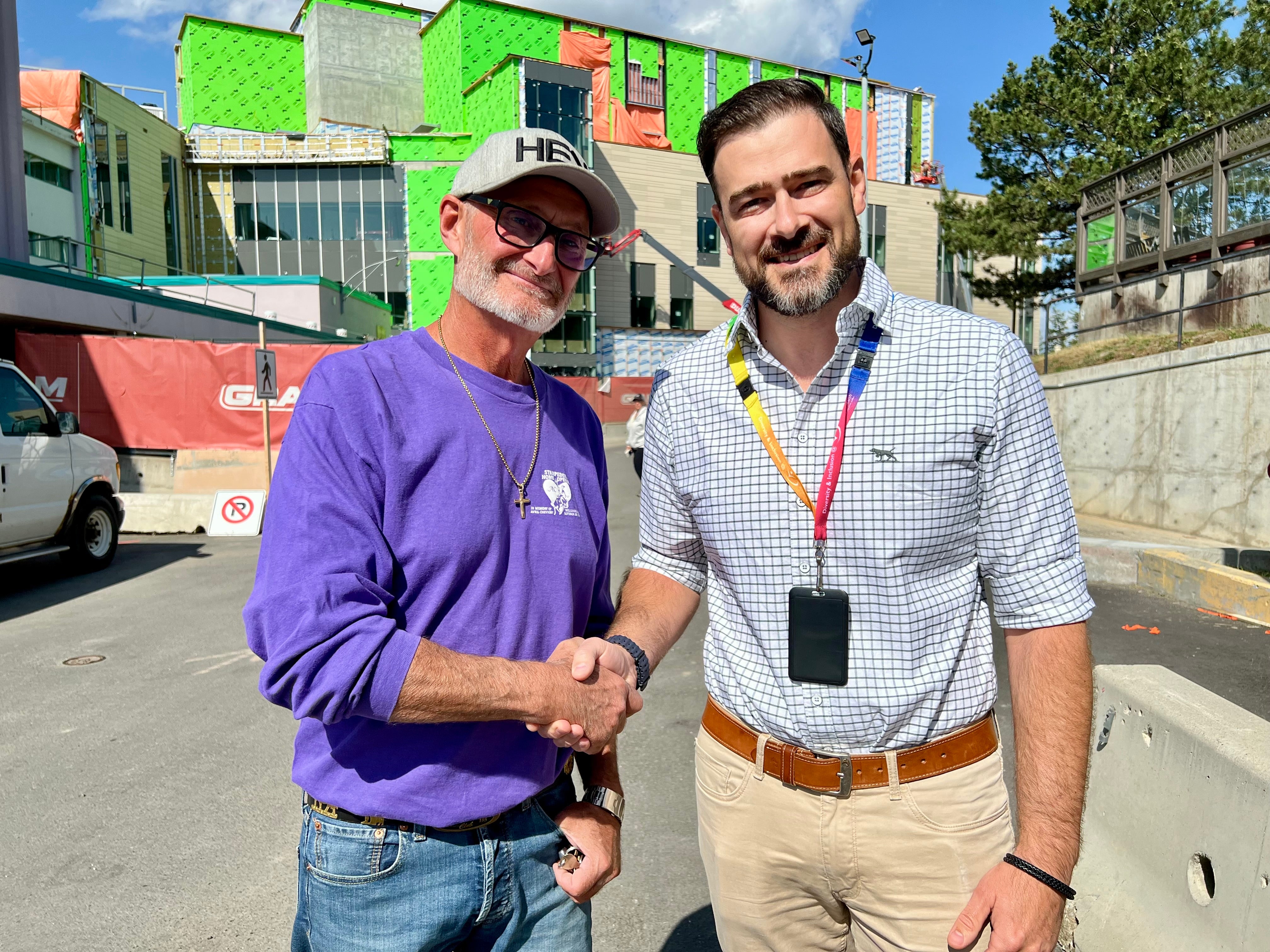 Two pepole, one in a purple shirt and one in business casual clothing, shakes hands in front of a building under construction.