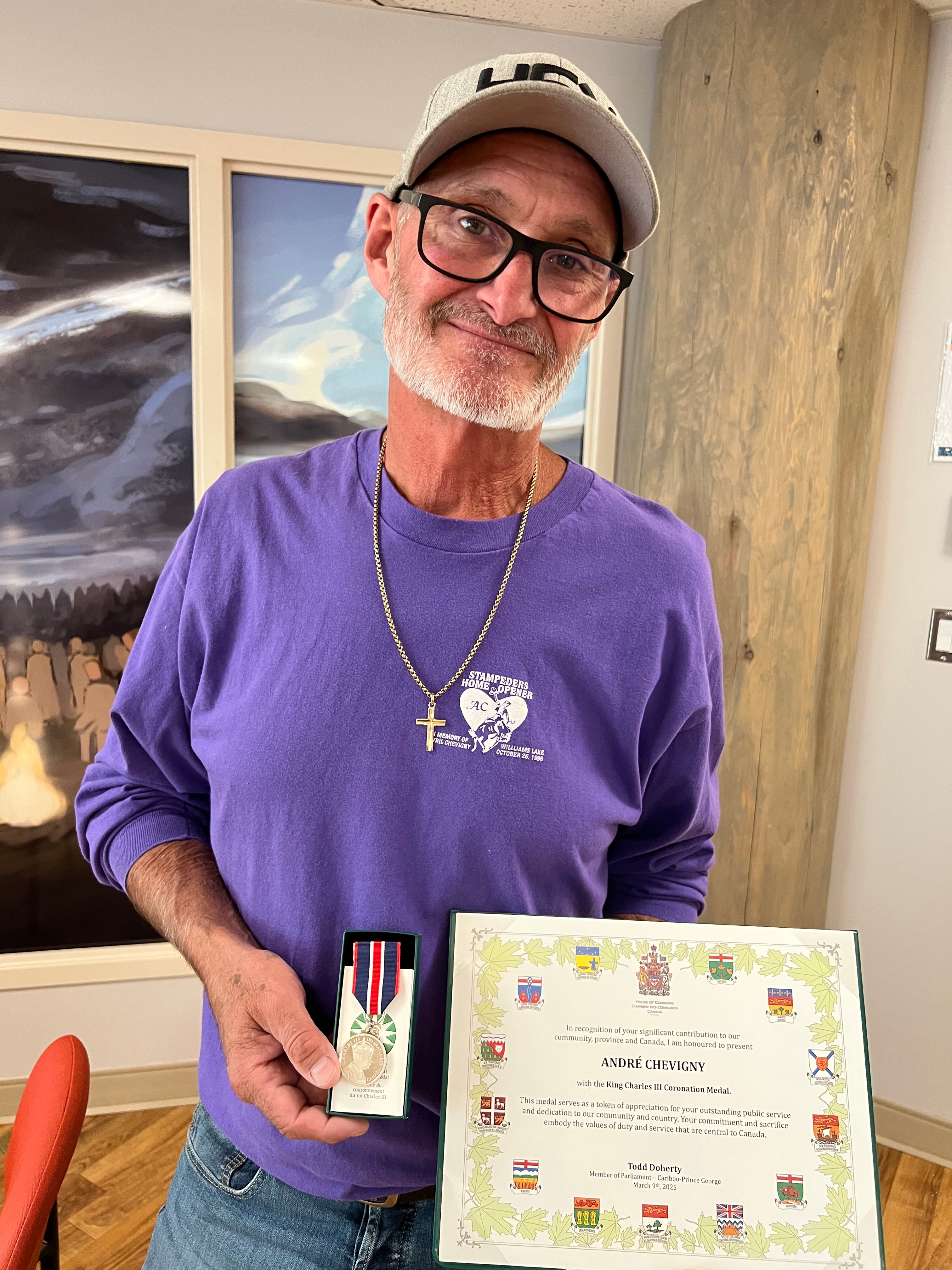 A person wearing a purple shirt, gold cross and ball cap holds a commemorative medal and certificate.