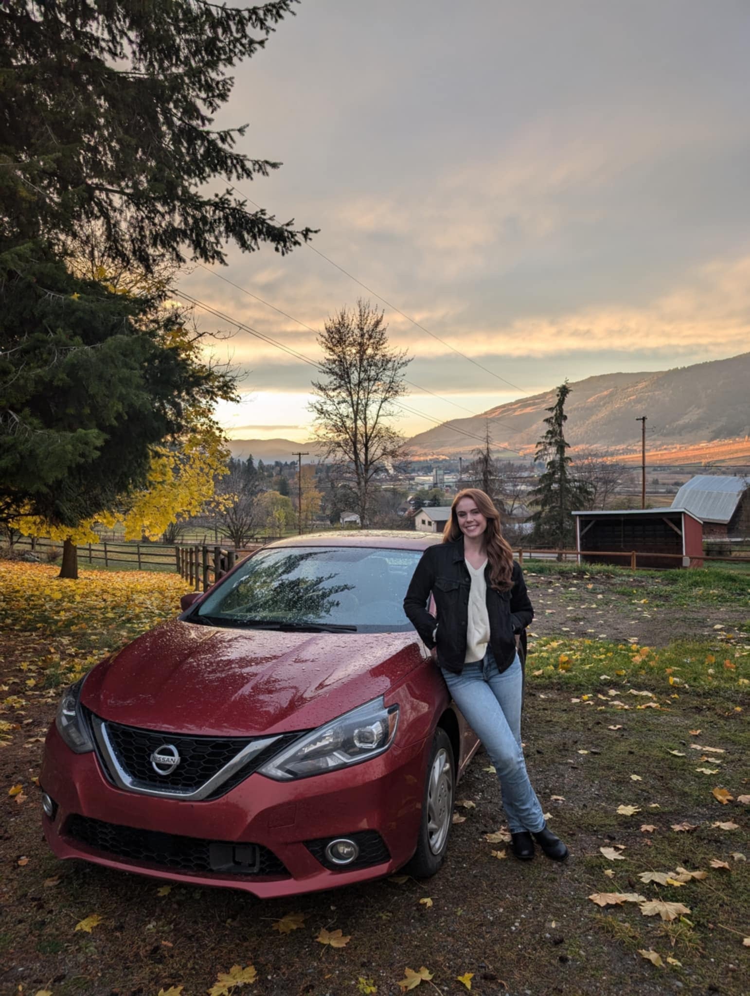 A casually dressed person leans on a red car outside on a fall day.