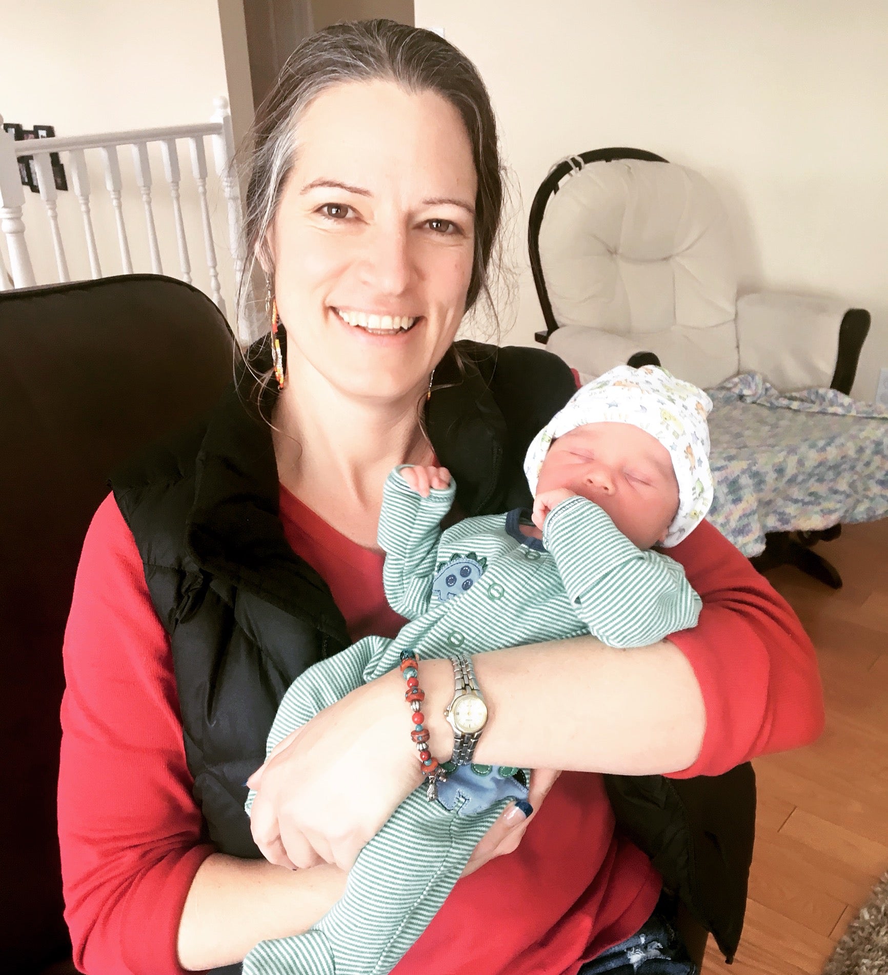 A person sitting in a chair holding a newborn baby swaddled in a white hat and blue jumper.