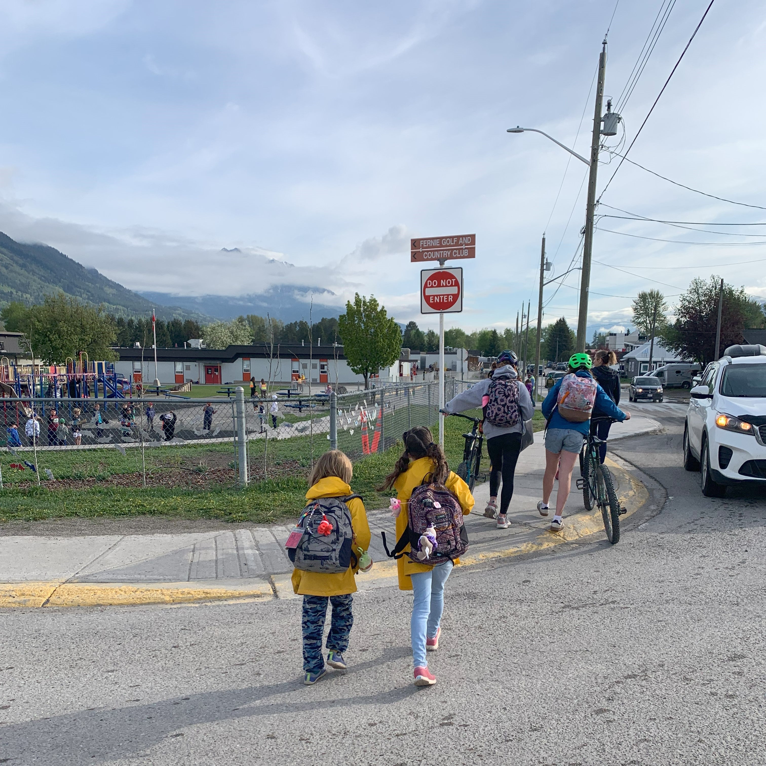 Two children in bright yellow rain jackets and carrying knapsacks cross near a do not enter sign near a school ground.