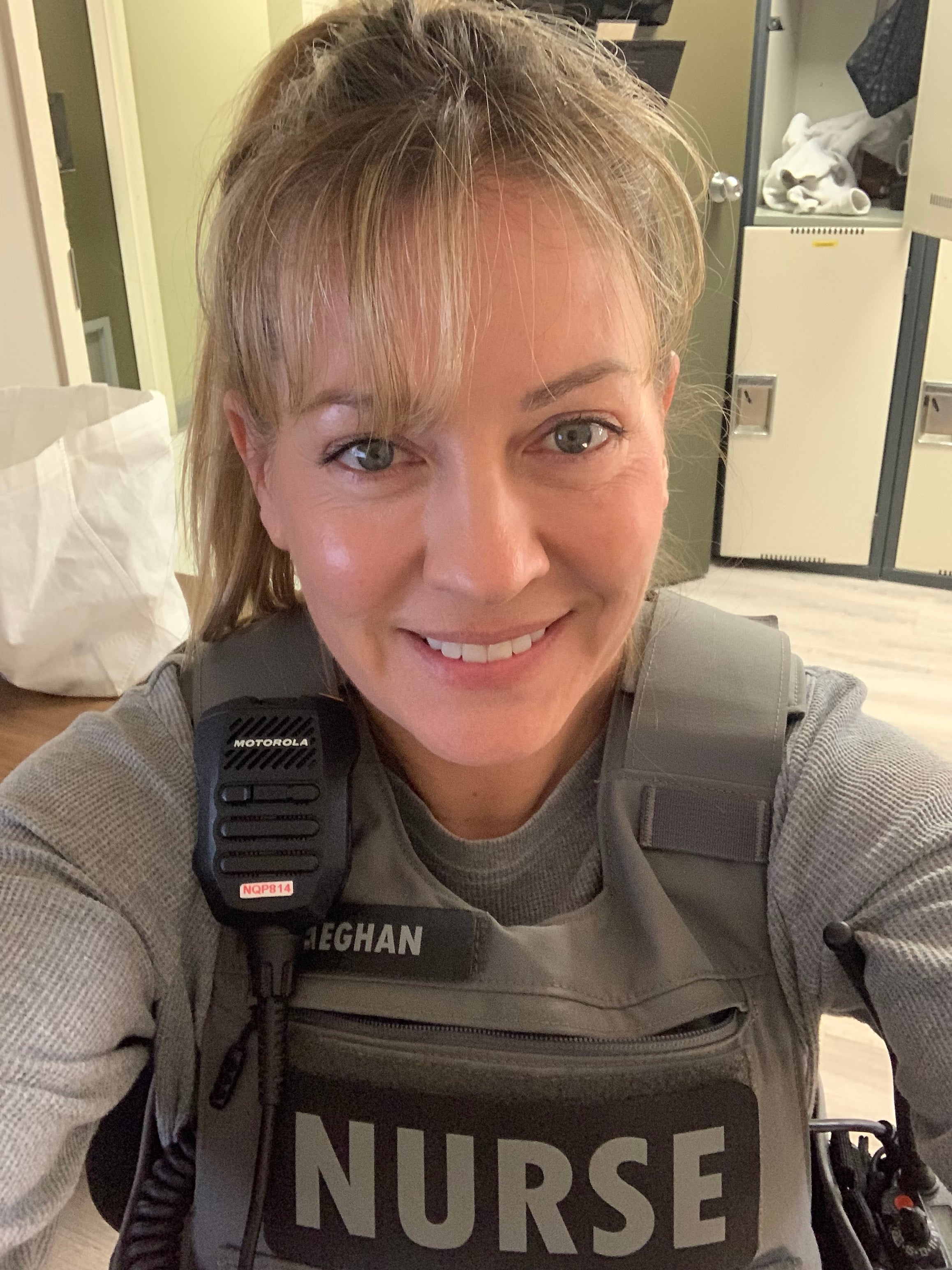 A nurse wearing a protective vest labelled “nurse,” smiling in a clinical setting.