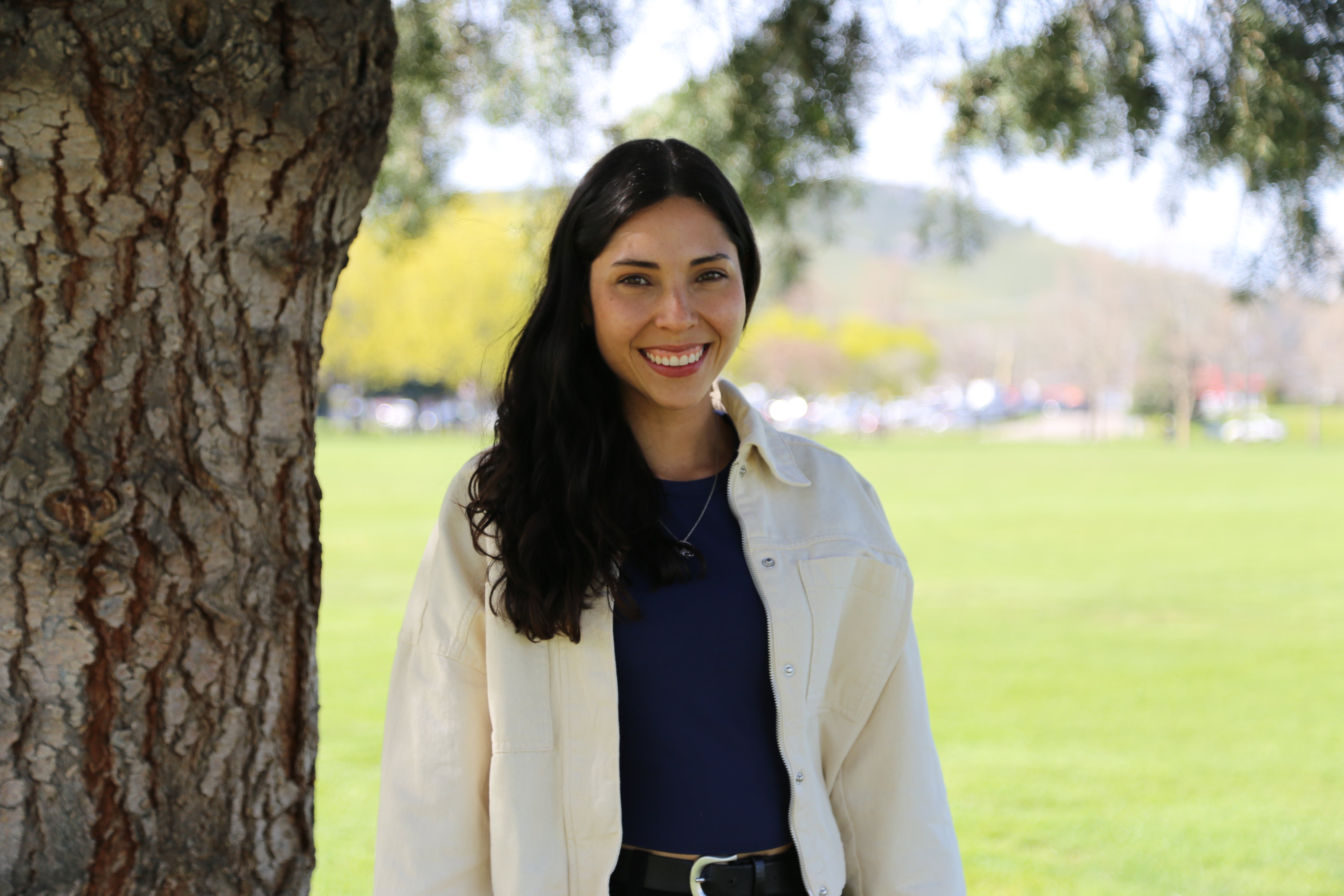 A smiling person with long dark hair wearing a white jacket and black shirt stands under a tree in a park.