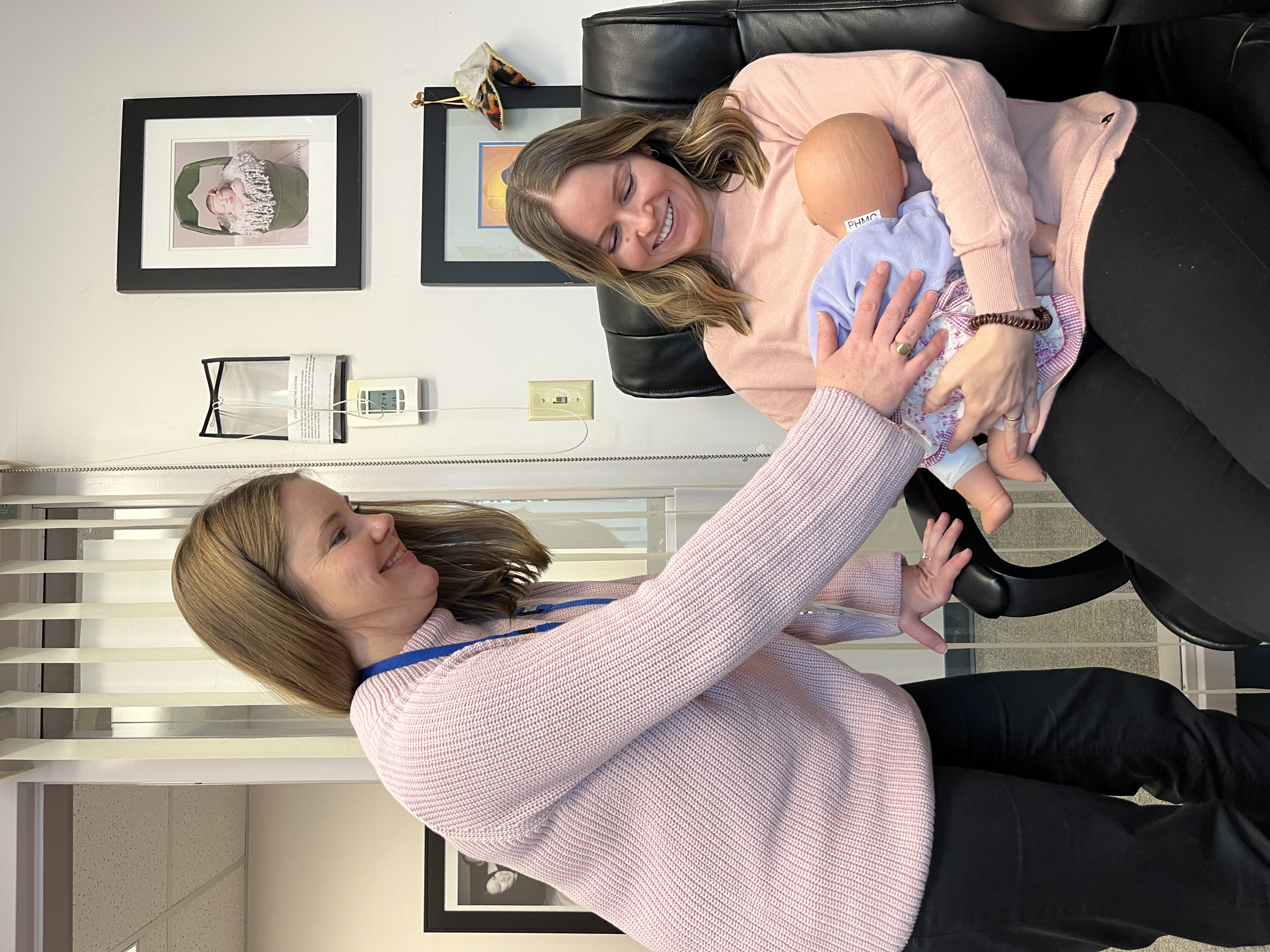 A person in a pink sweater with brown hair stands next to a person in a peach shirt sitting in a chair with a doll to demonstrate how to hold a baby during breastfeeding