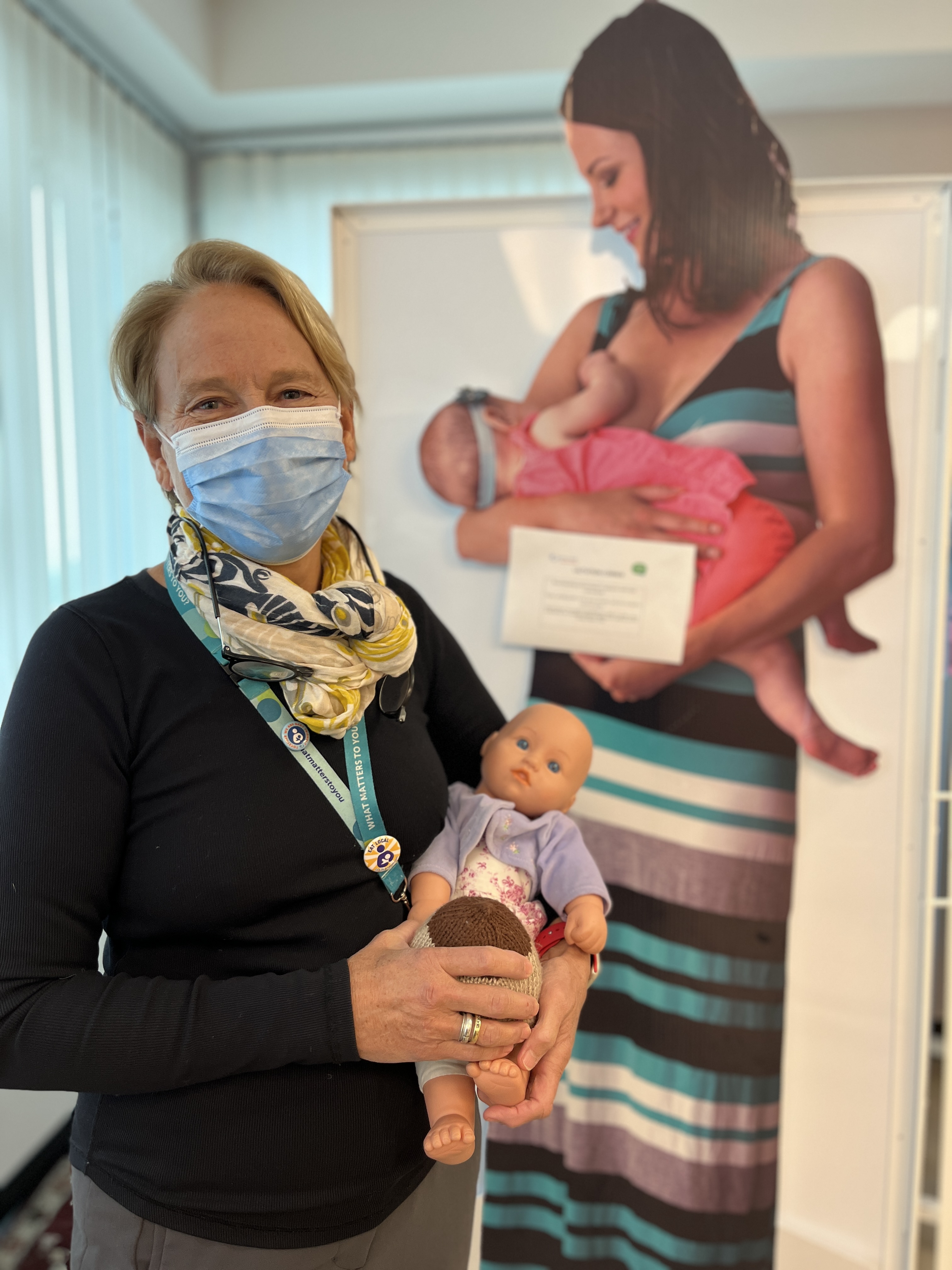 A person with short blonde hair wearing a black sweater, yellow scarf and a lanyard holds a baby doll in front of a poster display on breastfeeding