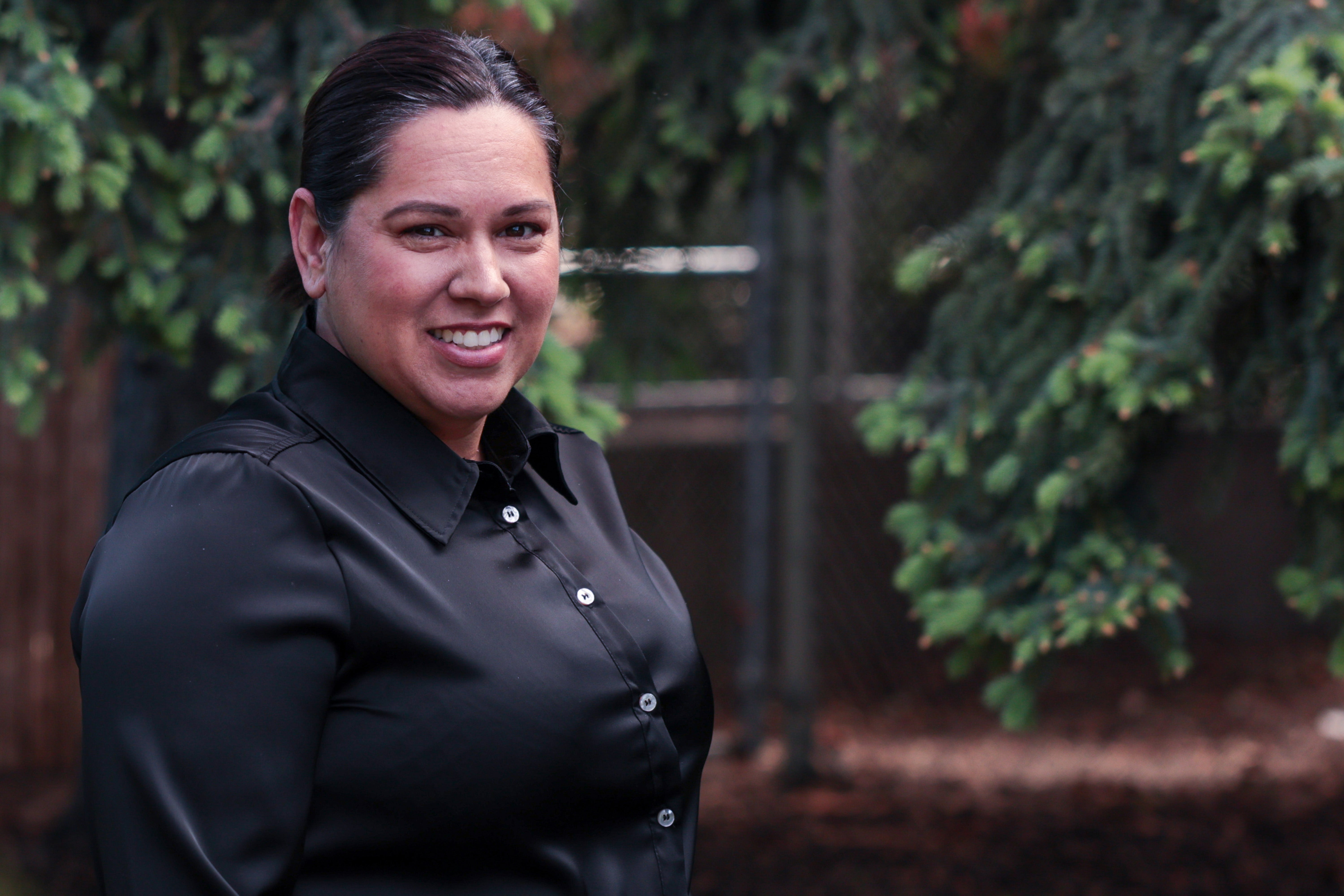 A smiling adult who has dark hair and is wearing a black shirt poses for a picture outside in front of tree branches 