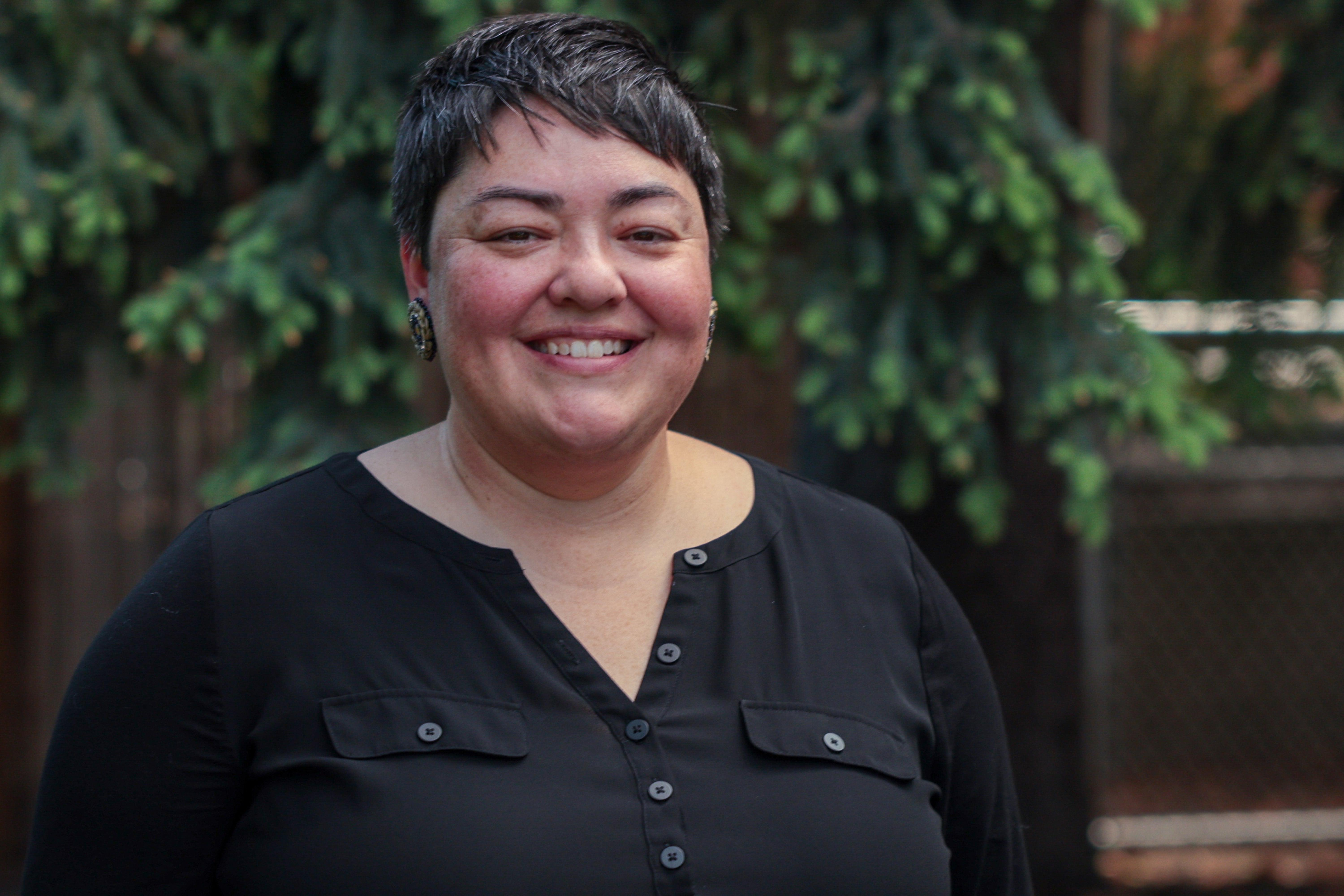 A smiling adult who has dark hair and is wearing a black shirt poses for a picture outside in front of tree branches 