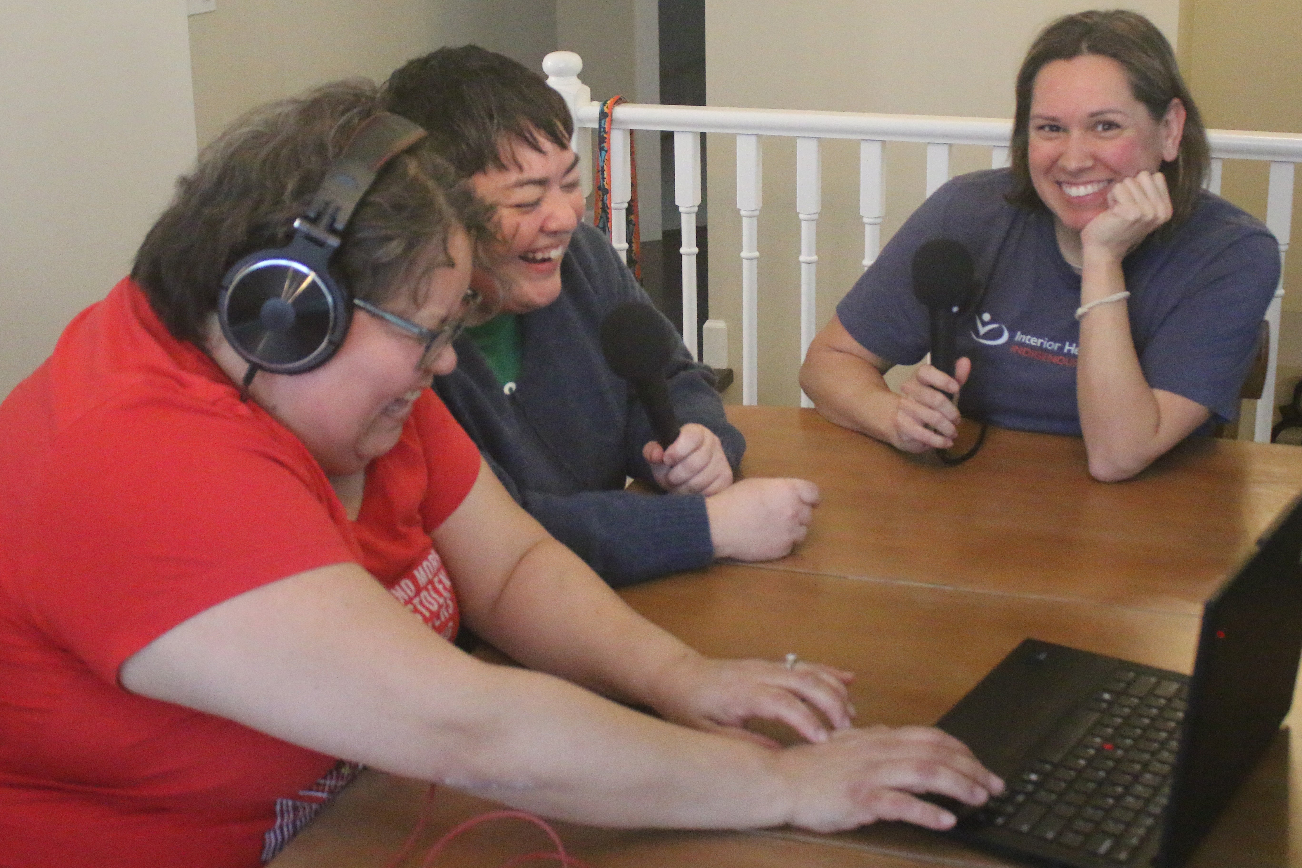 Three people laughing around a table, two of them holding microphones and another wearing headphones and interacting with a laptop.