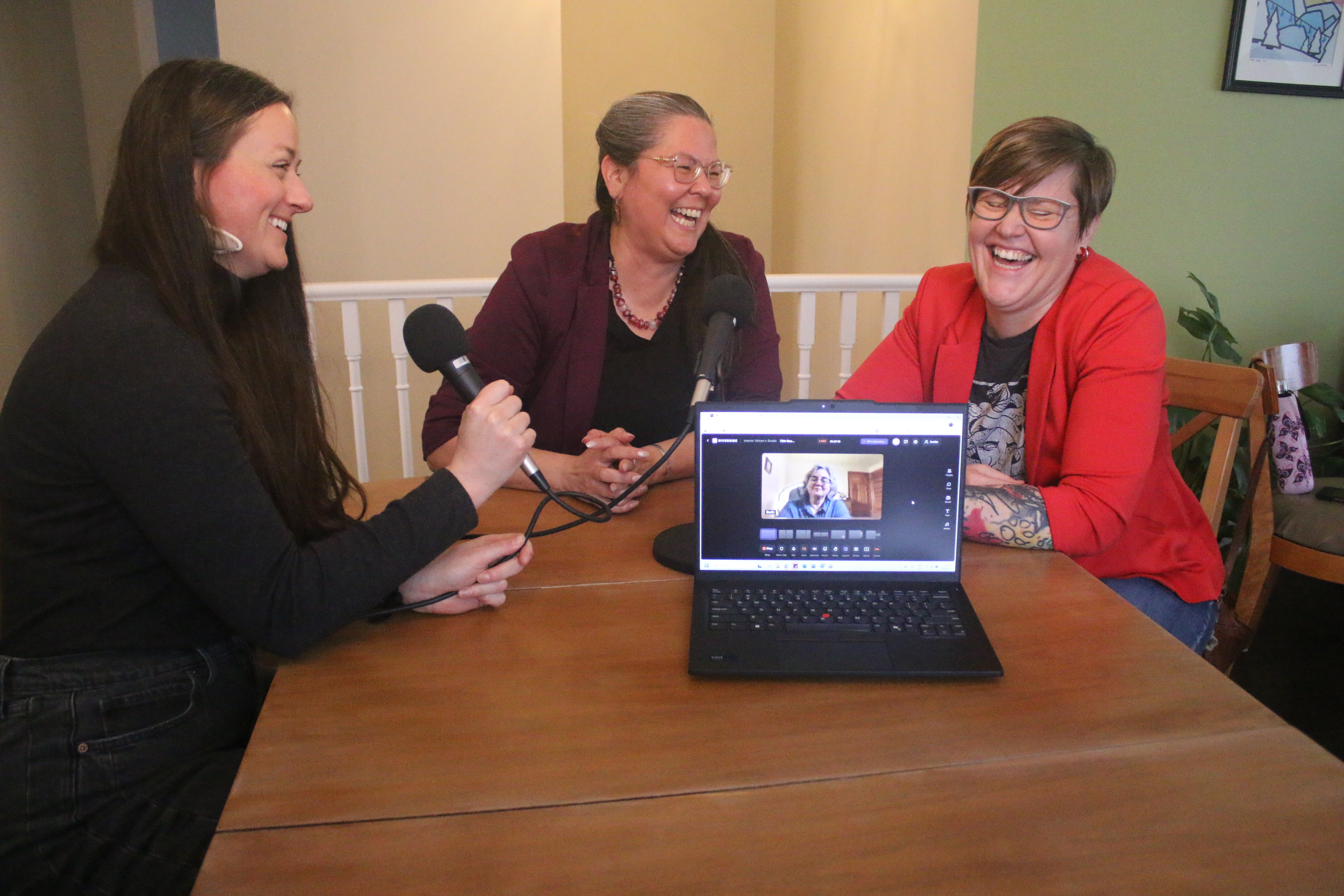 Three people laughing around a table that has two microphones set up and a laptop with a person connected virtually.