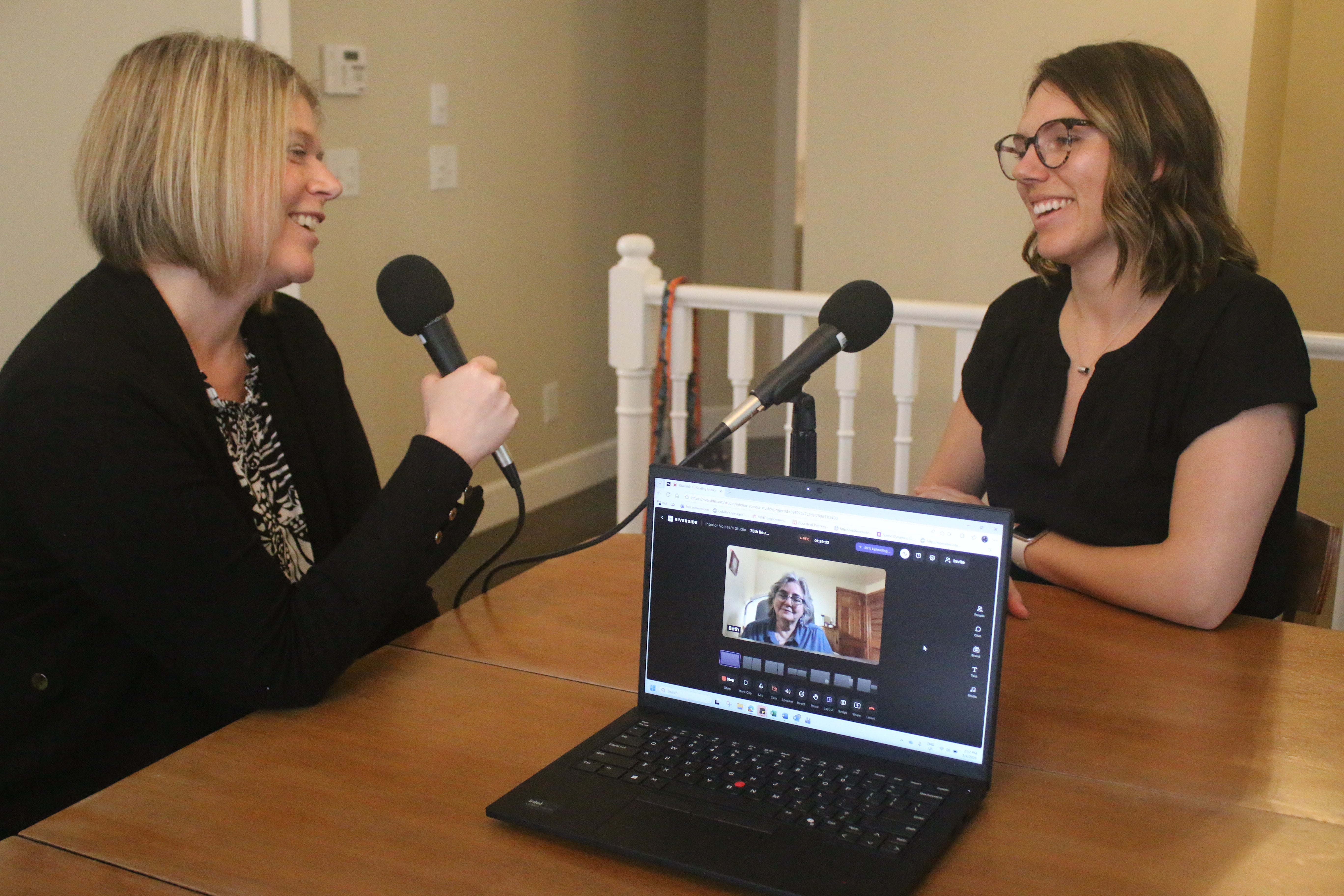 Two smiling people sitting across from each other speak into microphones, and a third person is connected virtually via a laptop on the table.