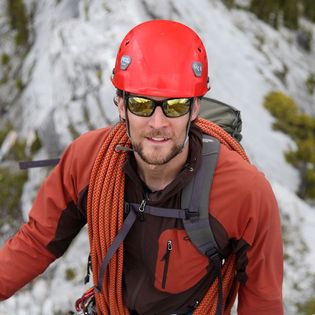 A man with a beard and wearing mirrored sunglasses, a red helmet, orange and black jacket, and climbing rope poses at the top of a rock face