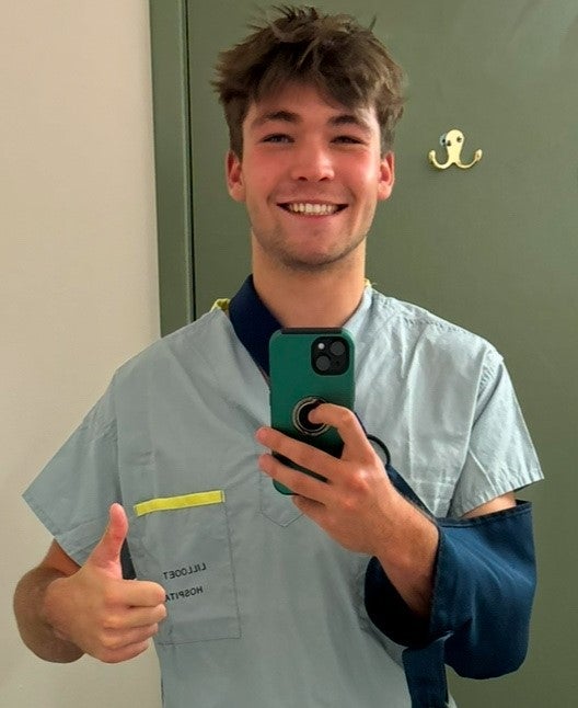 A young man with curly brown hair and light facial hair wearing green medical clothing and a heart pressure monitor gives a thumbs up as he takes a mirror selfie.