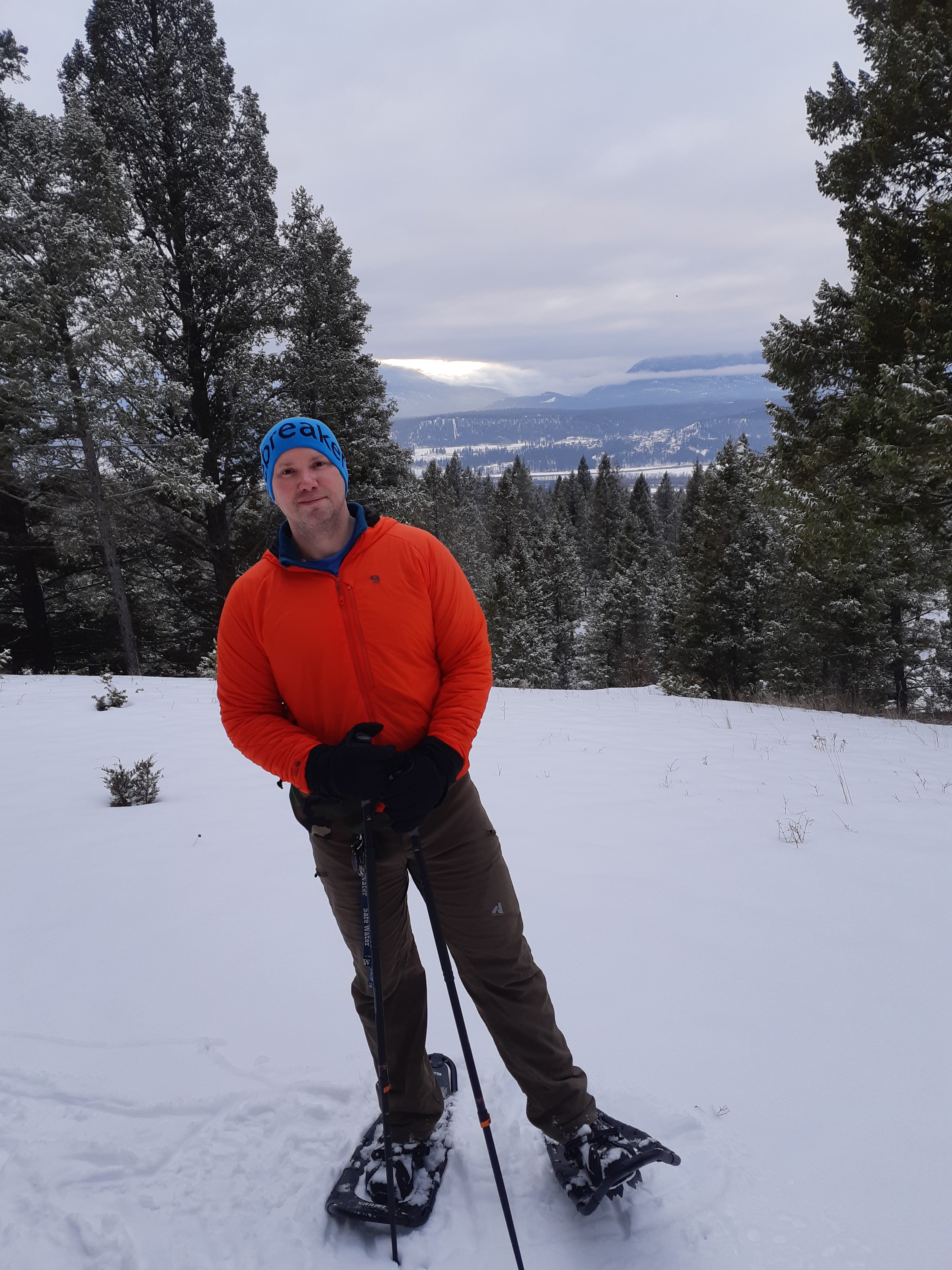 A man in a blue toque and red ski jacket poses with snowshoes and poles on a snowy trail with evergreens and mountains in the background