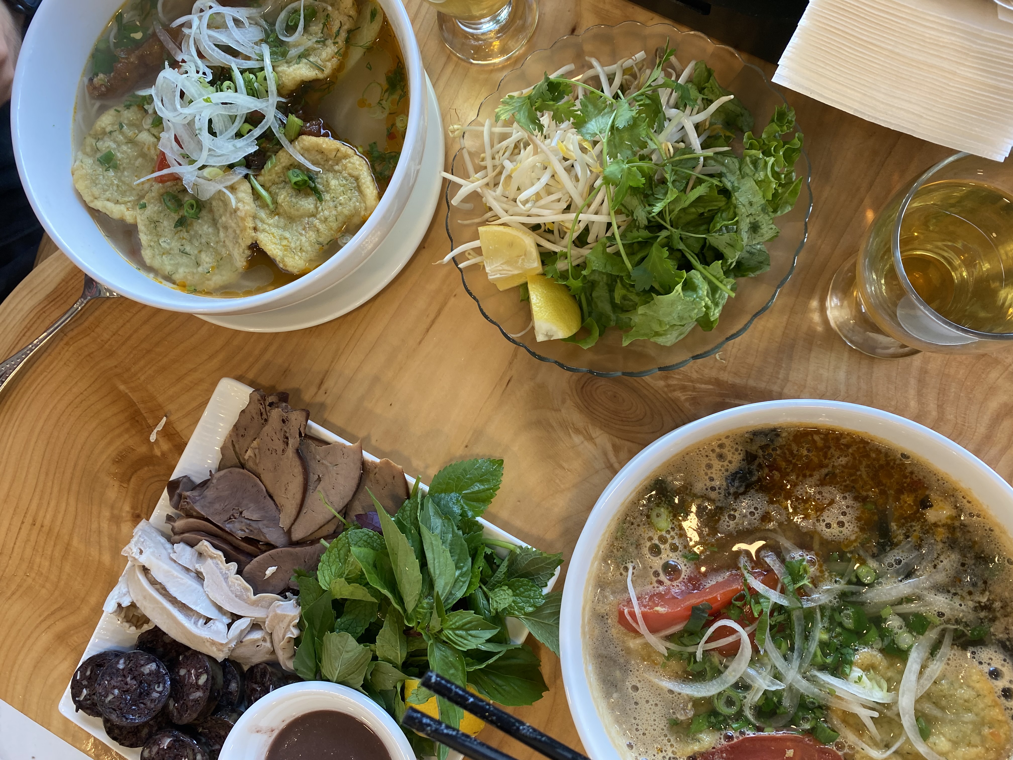 An assortment of food is on a table including vegetables, meats, noodle soups and drinks. 